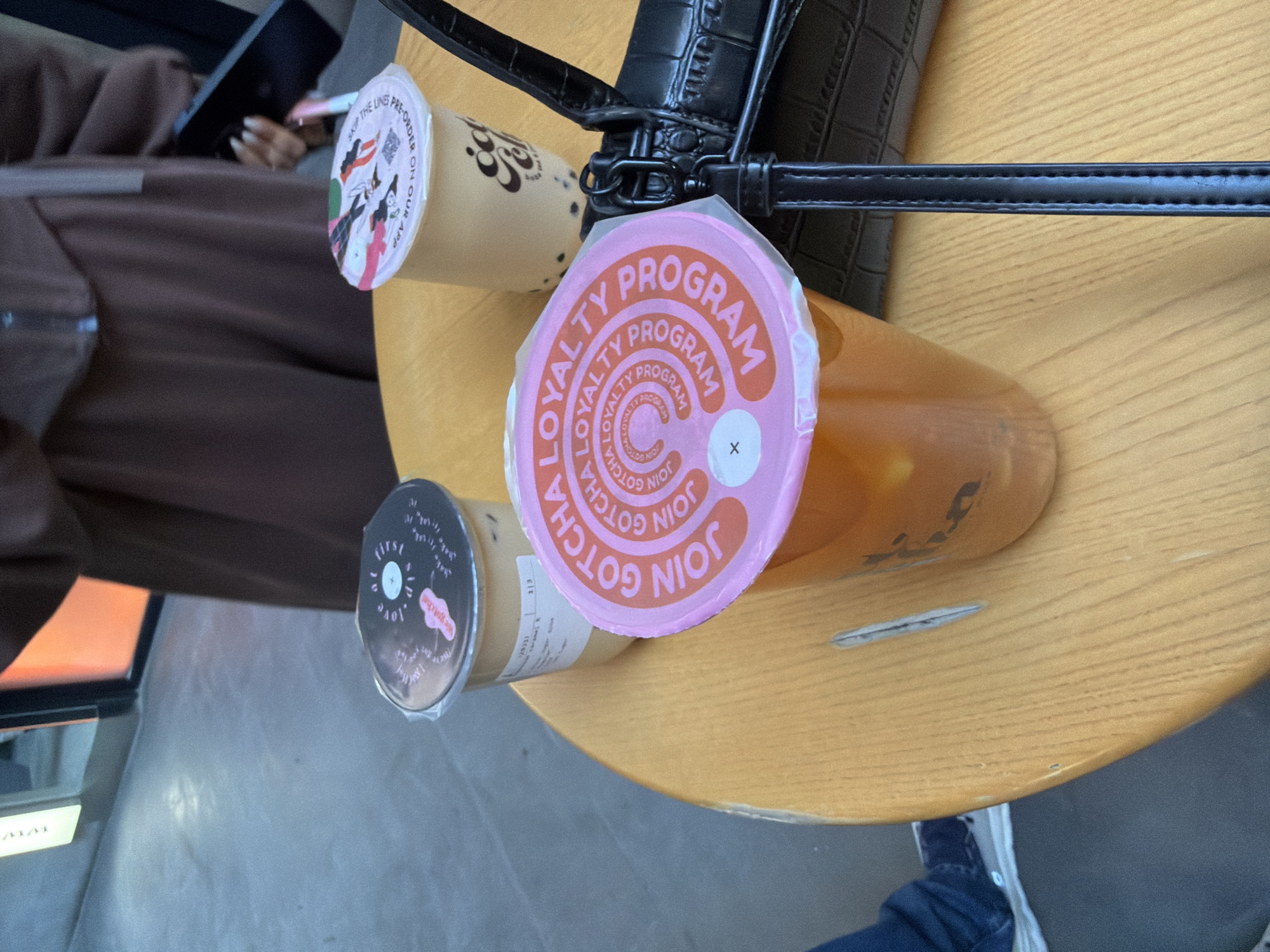 This image captures an indoor scene, likely a boba tea shop or cafe in Yerevan, Armenia. The central focus is on three plastic cups of boba/bubble tea, arranged on a light-colored wooden table. A black handbag with a textured surface and a dark strap is visible, resting beside the drinks.
The most prominent drink features a bright pink lid with circular text repeatedly stating "LOYALTY PROGRAM" and "JOIN GOTCHA," suggesting a promotion for the brand's customer rewards. A small 'X' mark is visible on this lid. The beverage inside this cup is a clear, amber-colored tea. Another light-colored drink (possibly milk tea) has a lid with illustrated characters and text that reads, "SKIP THE LINES PRE-ORDER ON OUR APP." A third, also light-colored, drink has a darker lid featuring the phrases "first love" and "We gotcha!", with a side label indicating "caramel."
Part of a person's leg, clad in blue jeans and a blue and white sneaker, is visible beneath the table. In the soft-focus background, another individual is partially seen, wearing a brown top. Their hands, with long manicured nails, are holding a black smartphone and a pen, suggesting they are engaged with the device. An orange glow from what appears to be a screen is also faintly visible in the background. The wooden table shows some minor wear, including a small scratch. The overall ambiance is casual and relaxed, indicative of patrons enjoying beverages and personal activities. The presence of the "Gotcha" branding ties into the provided location context of Yerevan, Armenia, as Gotcha Fresh Tea is an international chain.