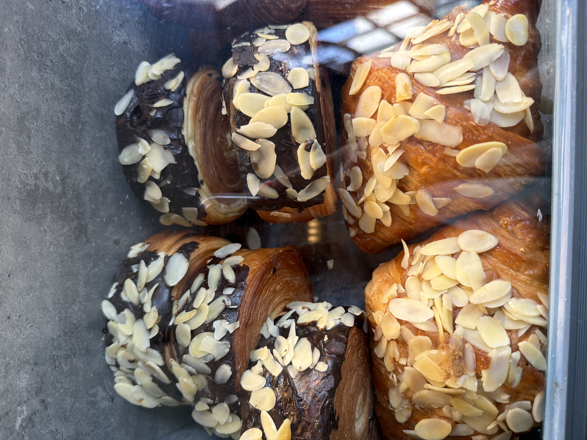 This image shows a close-up, eye-level view of several baked goods, likely croissants or pastries, displayed in a glass case. The pastries are topped with sliced almonds and some appear to be glazed with chocolate. They are arranged in layers, with the bottom layer showing golden-brown, flaky pastry and the top layers featuring the chocolate glaze and almonds. The background is a plain, dark gray textured surface, suggesting the interior of a bakery or display cabinet. The lighting highlights the textures and colors of the pastries. The image does not provide any information about the city of Đà Nẵng, Vietnam, or any people or actions.