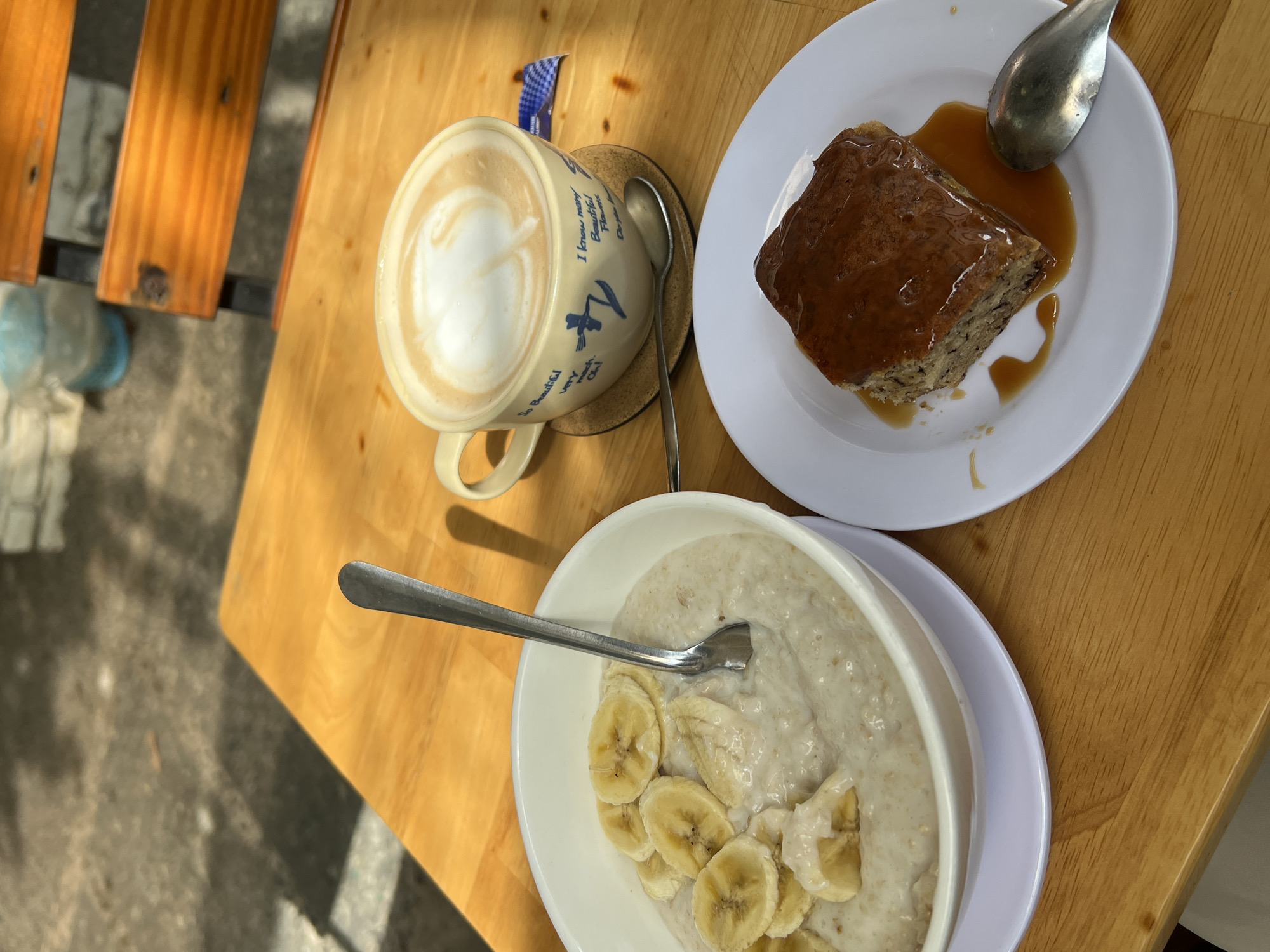 An overhead shot on a wooden table shows a cappuccino, a slice of banana cake with caramel sauce, and a bowl of oatmeal with banana slices. The mug of cappuccino is blue and white with writing, set on a small coaster. A spoon rests beside it. The cake slice has a dark brown caramel topping and sits on a small white plate with a spoon. The bowl of oatmeal, also on a white plate, is topped with banana slices and has a spoon in it. The scene is likely a cafe setting in Bac Nha Trang Ward, Vietnam, as this information was provided in the request.