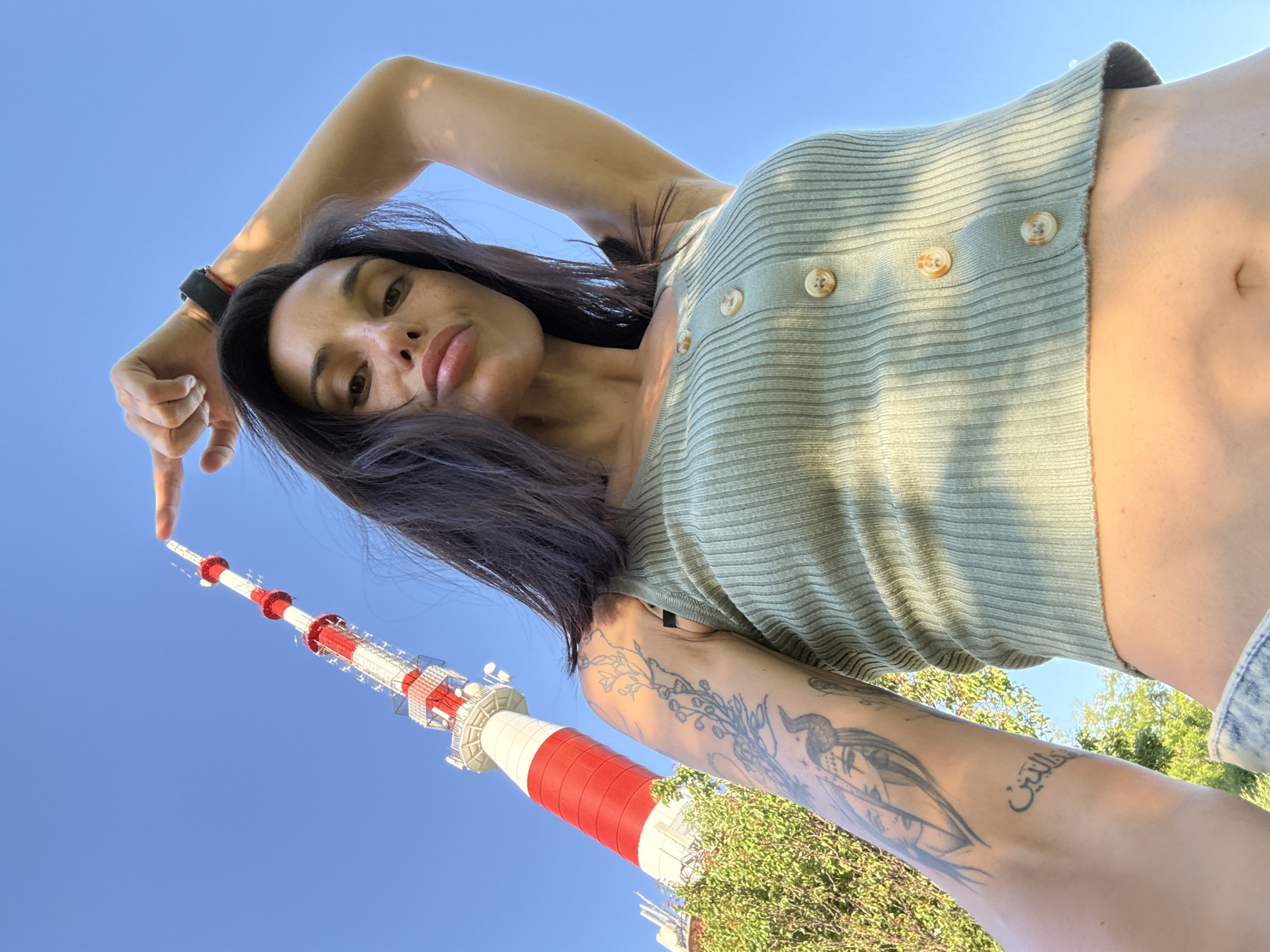 A low-angle shot shows a woman with dark hair and light brown skin standing outdoors, pointing with her right index finger toward the Pleven TV Tower, located in Pleven, Bulgaria. The woman is wearing a light green ribbed cardigan with brown buttons. She has tattoos on her left arm. The tower is red and white and is positioned against a clear blue sky. Green trees are visible near the base of the tower.