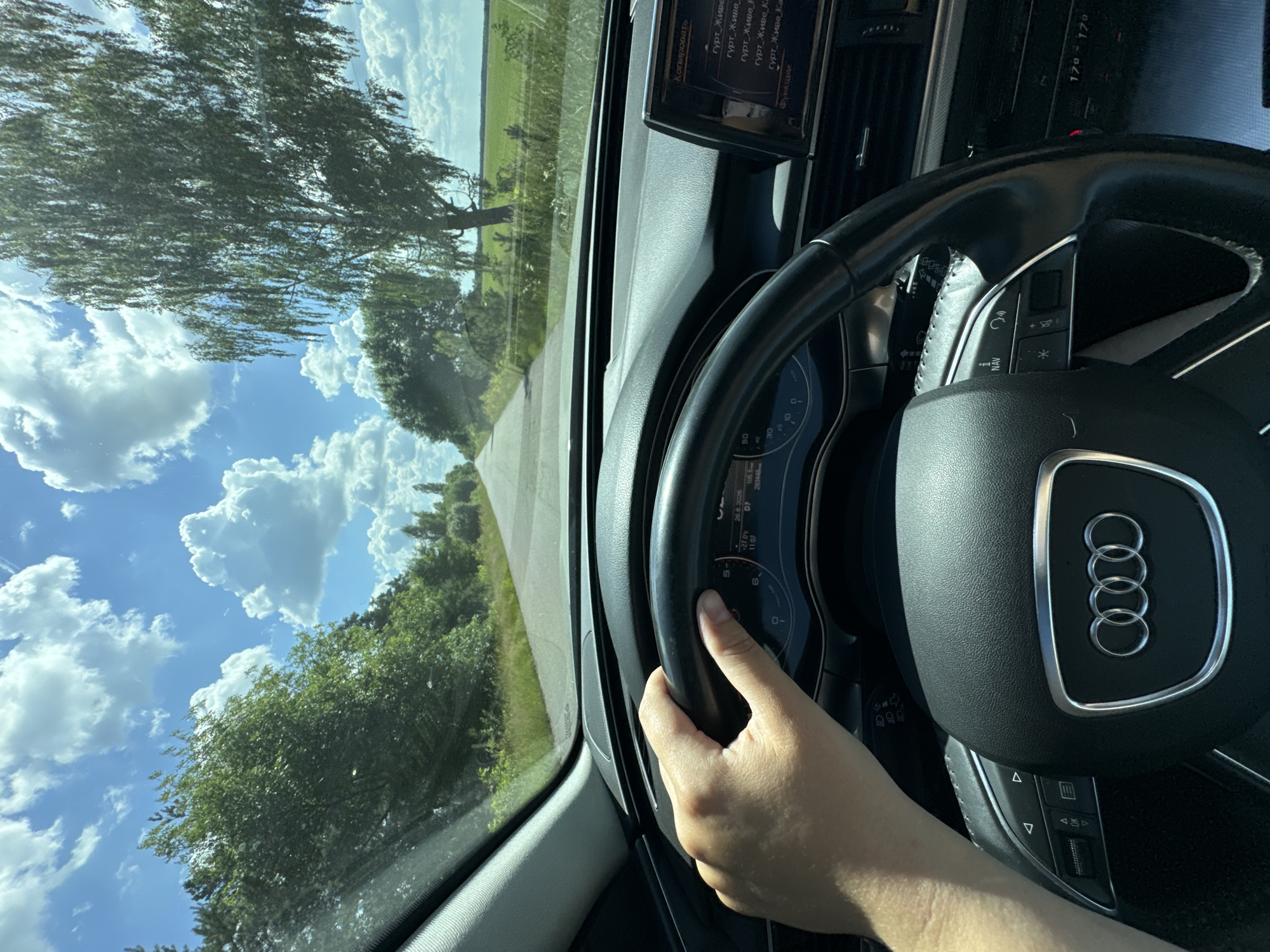 This image is taken from the perspective of someone driving an Audi. The driver's right hand is visible, gripping the steering wheel which features the Audi rings logo. Through the windshield, a road stretches into the distance, bordered by lush green trees and fields under a bright blue sky with fluffy white clouds. A large tree with thin leaves is prominent on the left side of the frame. The dashboard displays various indicators, including the time (11:07) and temperature (27.0℃), with some text in Cyrillic, suggesting a location in Eastern Europe. The scene appears to be set during the daytime, under clear and pleasant weather conditions, likely in a rural or semi-rural area.