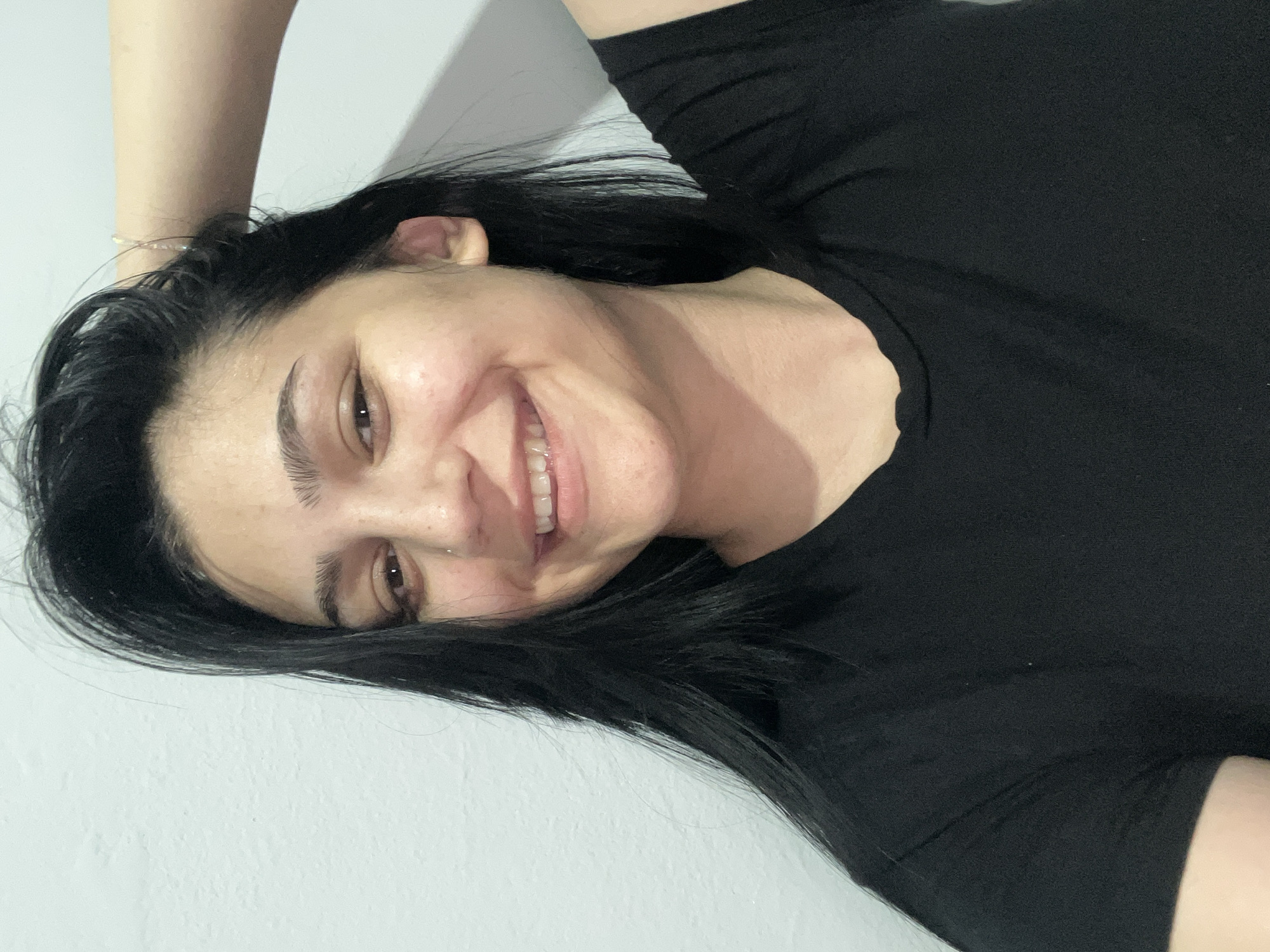 This is a close-up, high-angle shot of a young woman lying on her back, with her head resting on her arm. She has long, dark hair that falls around her face and shoulders. Her eyes are open and she is smiling, revealing her teeth. She is wearing a black t-shirt. The background is a plain white wall. The lighting suggests an indoor setting, likely during the daytime, though no specific time of day or weather conditions can be determined. The woman's expression conveys happiness and relaxation. There is no visible text in the image.
