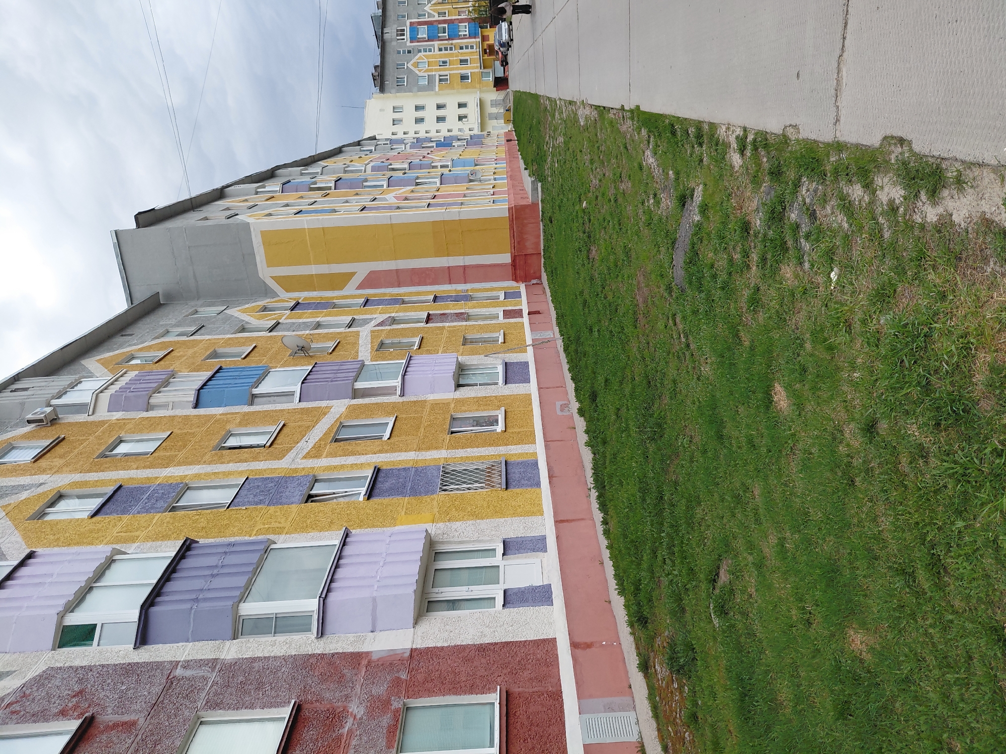 The image depicts a colorful apartment building in Nadym, Russia. The building has multiple stories and is painted with horizontal bands of various colors, including yellow, purple, blue, and red. The facade has numerous windows with some having awnings of matching colors. A narrow strip of green grass separates the building from a concrete sidewalk. In the background, other apartment buildings can be seen. Overhead power lines are visible against a cloudy sky. The image has a tilted perspective, giving an upward view of the building.