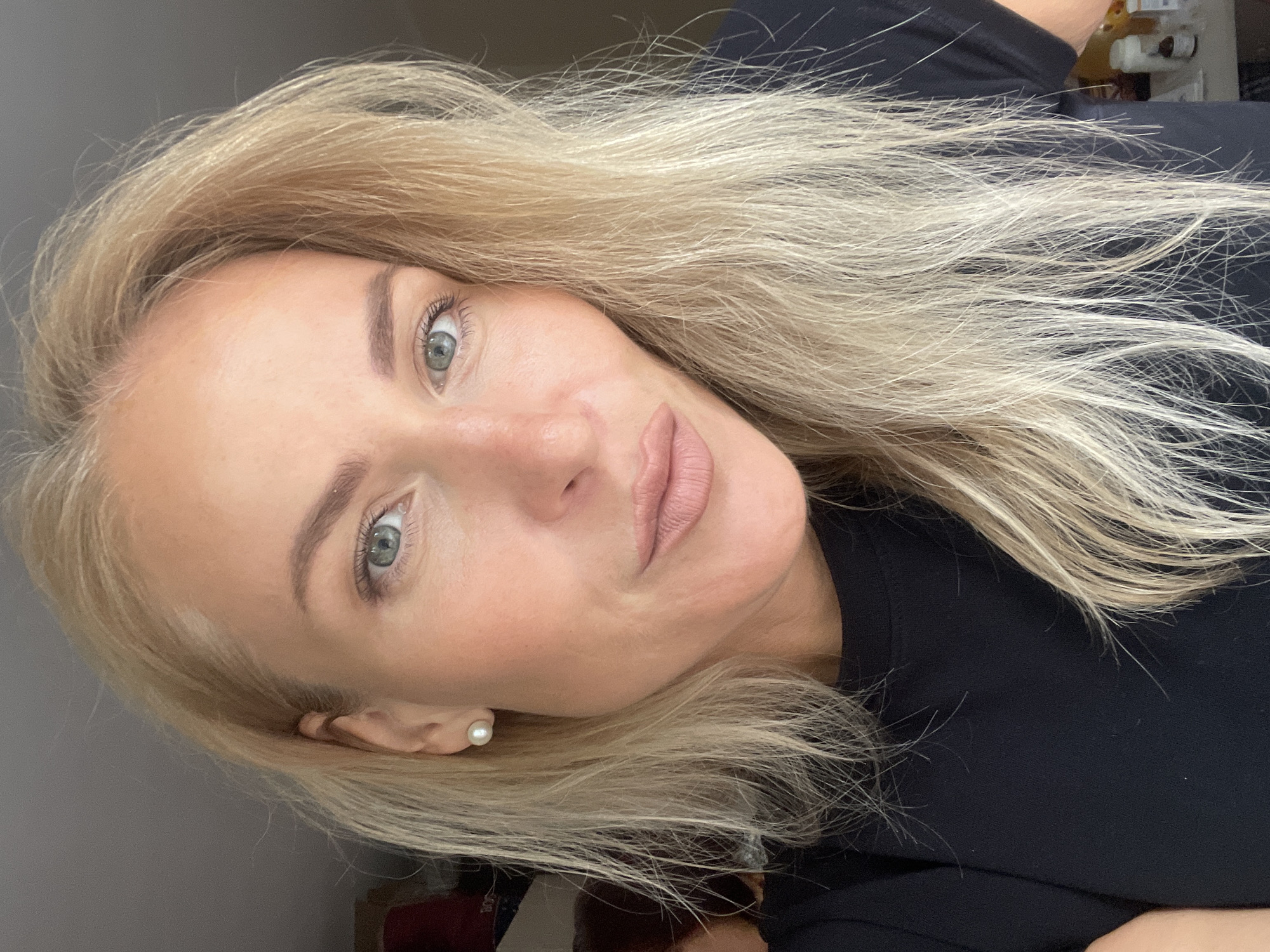 A portrait of a blonde-haired woman with light skin and blue-green eyes, wearing a black shirt and pearl earrings. The location is indeterminable from the image.
