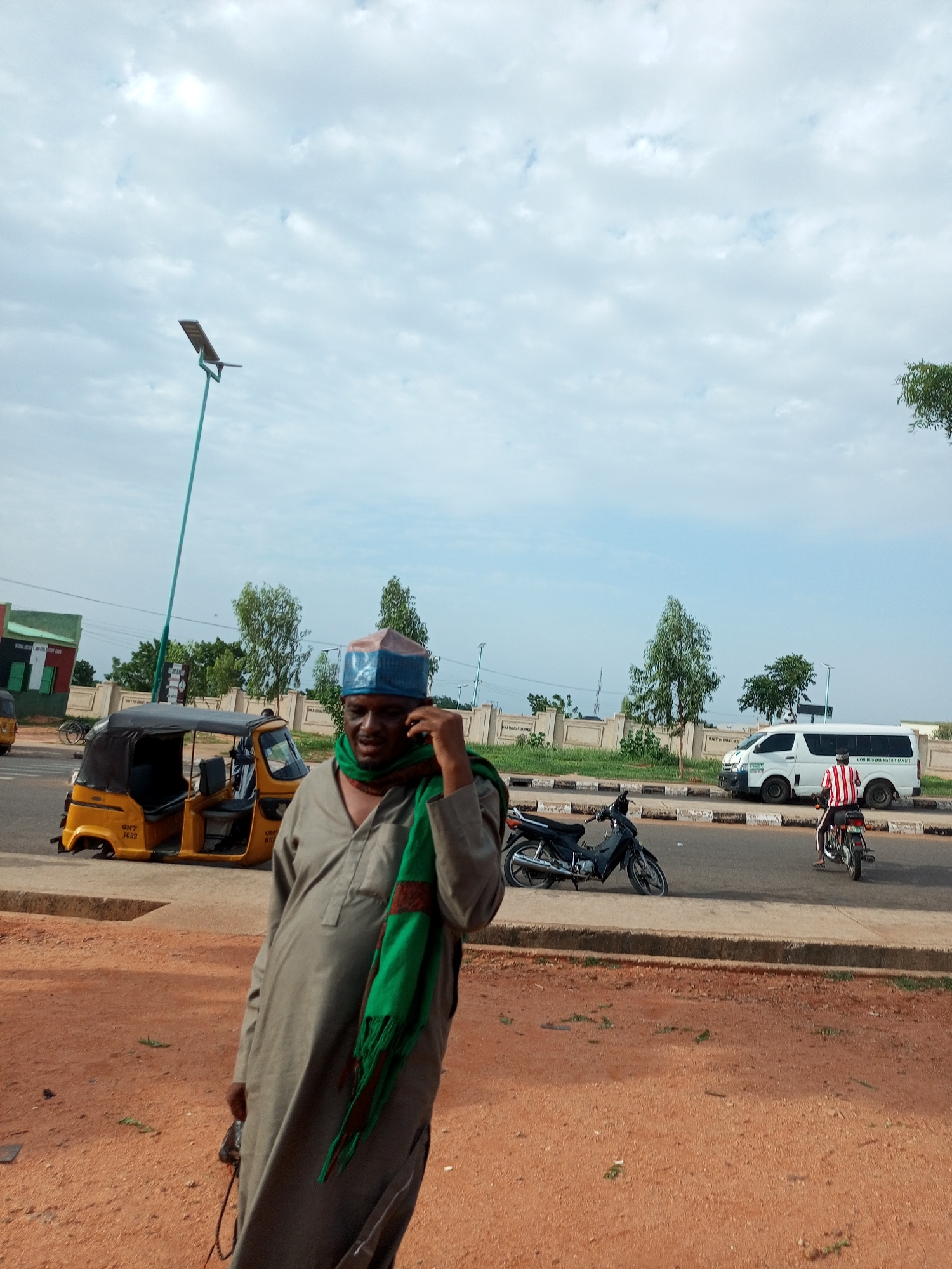 A man stands on a dirt road in Gombe, Nigeria, holding a phone to his ear. He wears a grey robe and a green scarf, with a blue and red cap. To his left, a yellow auto-rickshaw is parked. In the background, a white van and two motorcycles are on the road. The sky is cloudy.