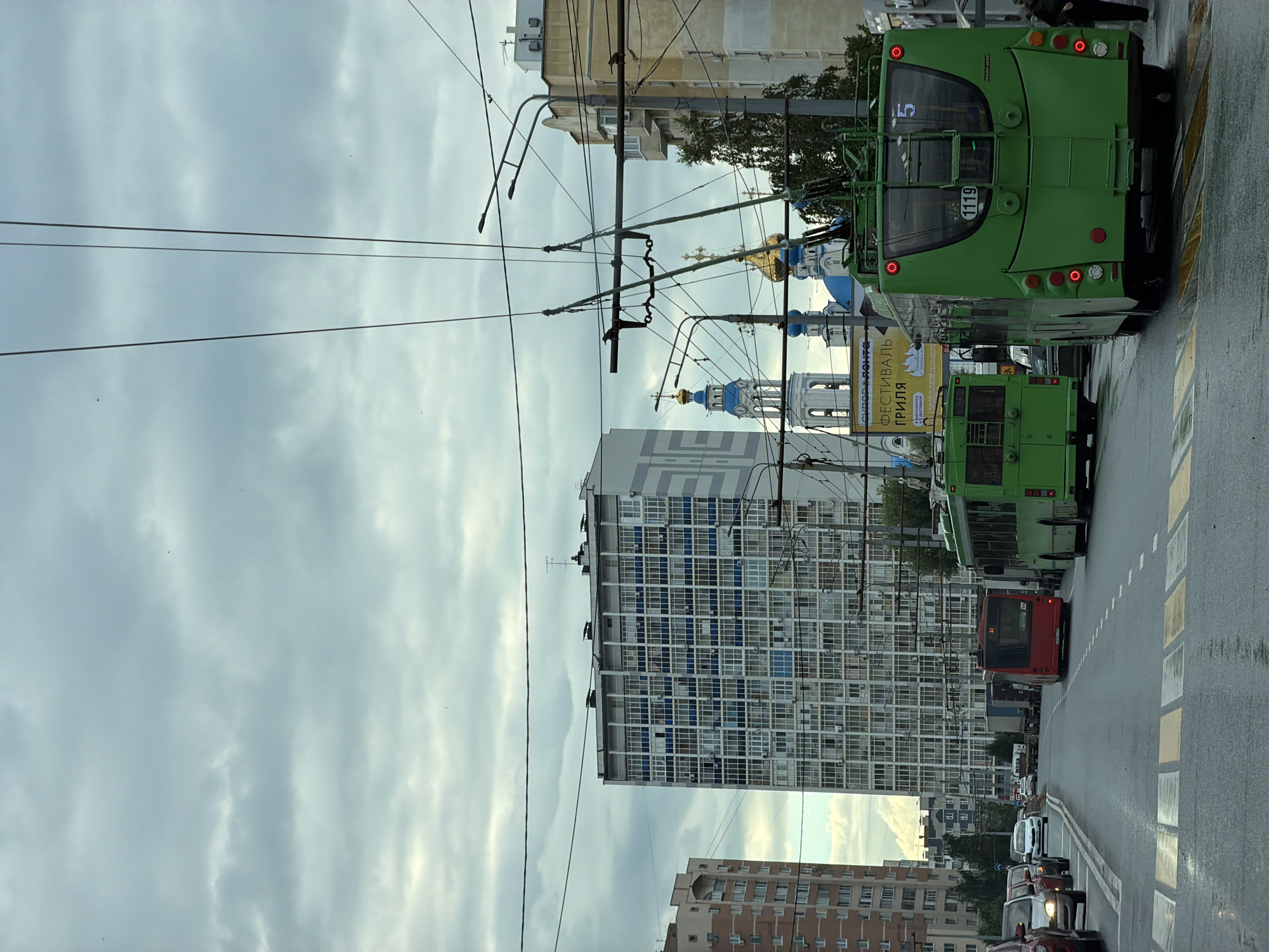 An overcast day in an urban setting. The street is wet, suggesting recent rain. Multiple lanes of traffic are visible, with several vehicles including red and green buses, cars, and trucks. Overhead power lines crisscross the sky, indicating a trolleybus system. In the background, a tall, modern apartment building with many windows dominates the skyline, while to the left, another apartment building with a red-toned facade is partially visible. Further in the distance, several church domes, one with a golden dome and another with blue domes, are prominent features of the cityscape. A crosswalk is in the foreground.