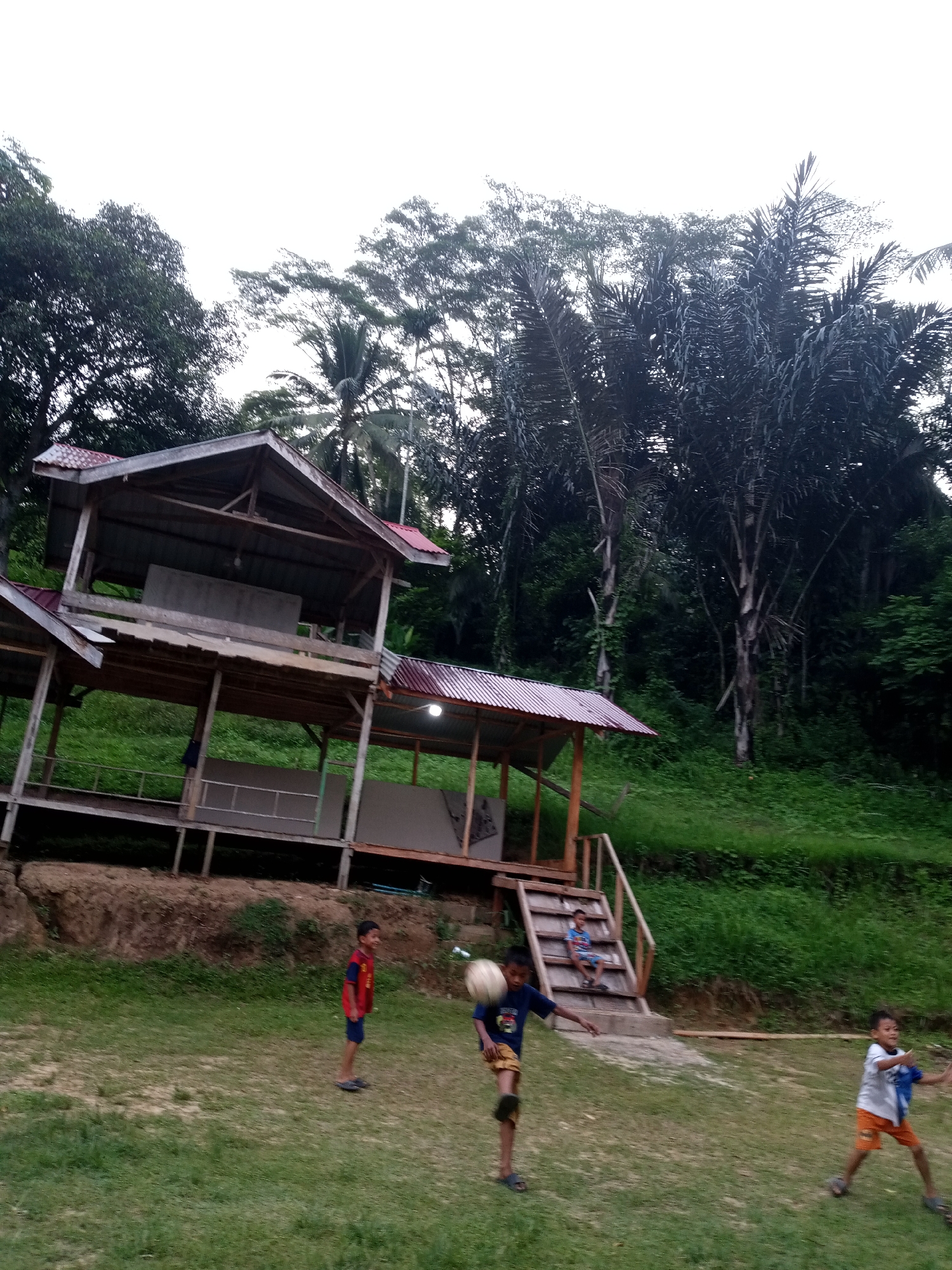 The image depicts a natural outdoor setting in the city of Lhokseumawe, Indonesia. In the foreground, three young boys are engaged in playing with a ball on a grassy field. One boy, wearing a red and blue shirt and dark shorts, stands to the left, looking towards the ball. Another boy, in a dark blue shirt and khaki shorts, is in the center, mid-kick, with the ball airborne in front of him. To the right, a third boy, in a white and blue shirt and orange shorts, is actively participating, his arms extended as if to catch or pass.
Behind the children, a wooden structure with a corrugated metal roof is visible. This structure appears to be elevated and has multiple levels and open-air sections, suggesting it might be a pavilion or shelter. Stairs lead up to one of the levels where another young boy, wearing a blue and white striped shirt, is seated. The structure is built on a slight incline, with a grassy embankment behind it.
The background is dominated by dense foliage and tall trees, indicative of a tropical environment. The sky above is overcast and bright white. The overall scene suggests a casual, recreational activity taking place in a semi-rural or park-like area within Lhokseumawe.
