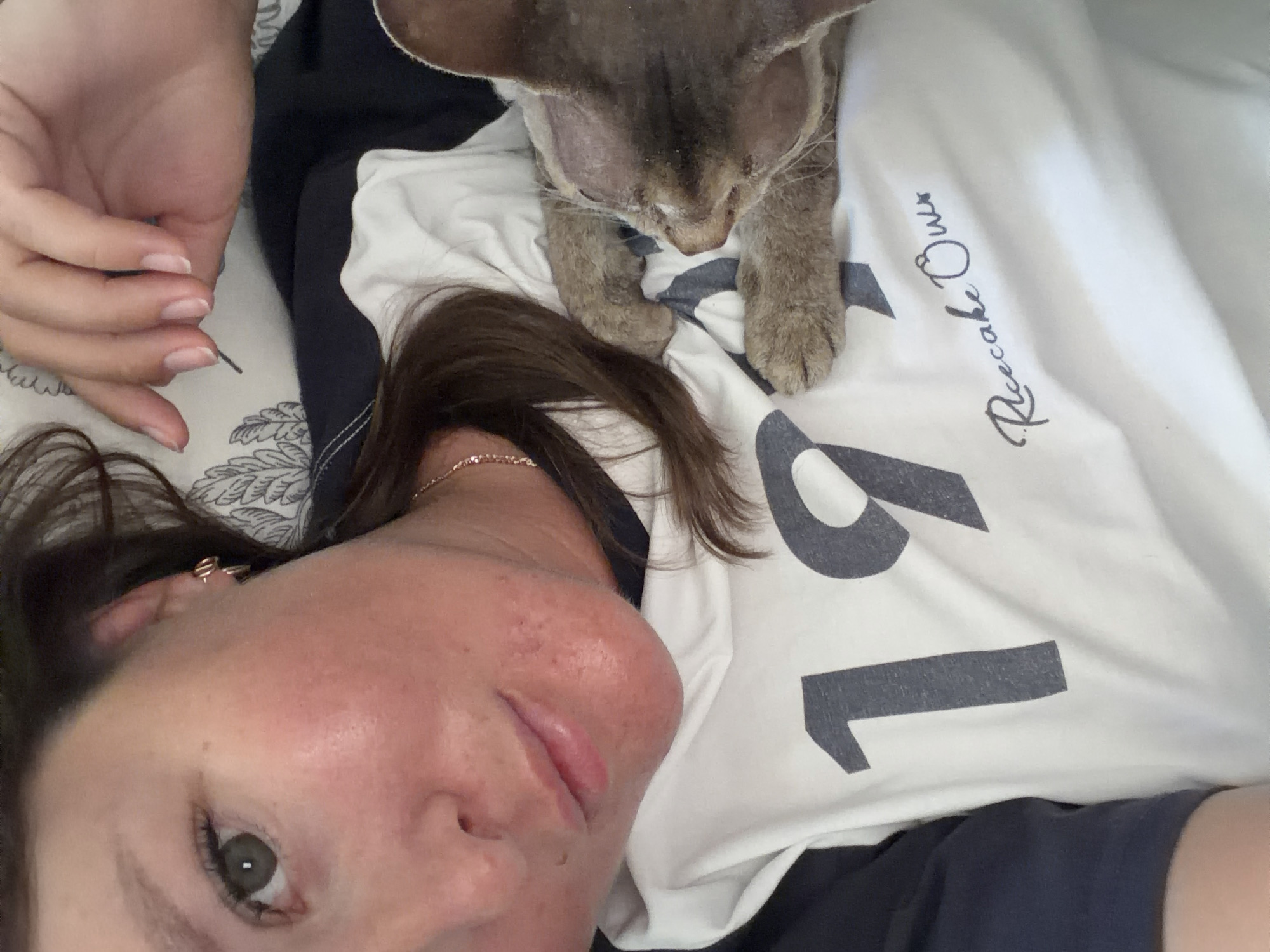 A woman is lying on her back, wearing a white t-shirt with black lettering. A cat is perched on her chest, with its paws resting on her t-shirt. The woman is facing upwards, with her face visible. The scene is indoors, possibly in a bedroom. The specific location in Chaikovsky, Russia, cannot be determined from the image.