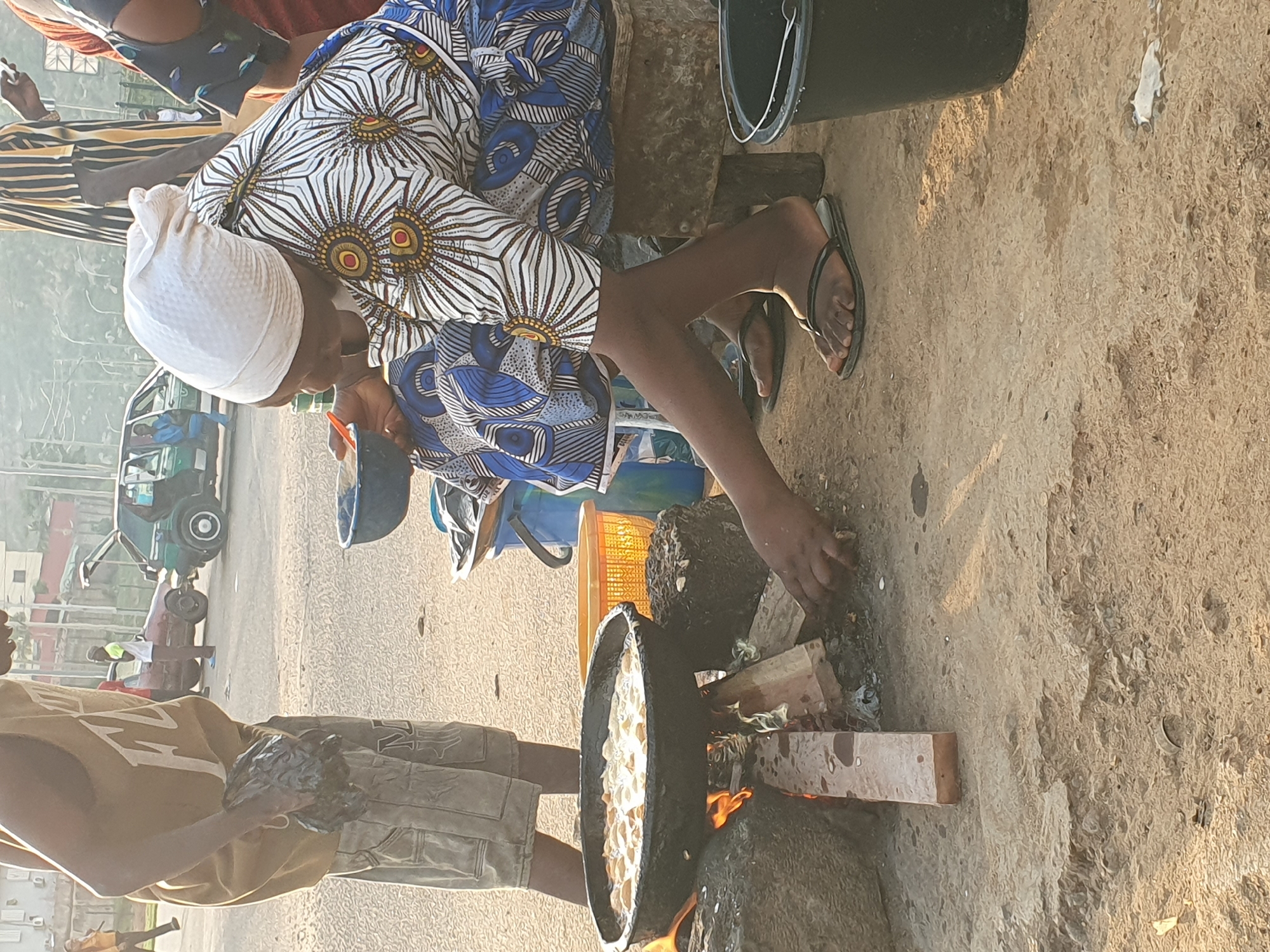 A woman in a patterned dress and white head covering is sitting near a small fire and cooking in a large, black pan. She is positioned next to the fire with her hand reaching into the pan. A person wearing a yellow shirt and grey shorts stands nearby. The ground is sandy. In the background, there is a vehicle and other individuals can be seen. The image appears to have been taken outdoors in the city of Abuja, Nigeria.