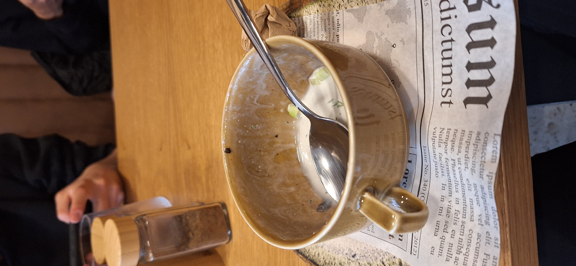 The scene depicts an indoor dining setting on a light-colored wooden table in Plovdiv, Bulgaria. In the foreground, a light brown ceramic bowl with a handle contains the residual white liquid of a consumed dish, likely a yogurt-based soup, with small green pieces resembling cucumber and dill. A silver spoon rests inside the bowl. The bowl is positioned on a sheet of paper designed to mimic a newspaper, featuring placeholder Latin text such as 'Lorem ipsum' and 'Issue No. 340 (C)', which is itself placed on a circular, mottled brown placemat. To the left of the bowl, two clear glass spice shakers with light wooden caps are visible; one contains dark granular seasoning. In the blurry background, the upper torsos of at least two individuals are present. One person on the left is visible holding a mobile device with both hands. The background consists of a warm brown, indistinct surface, possibly a wall or booth.