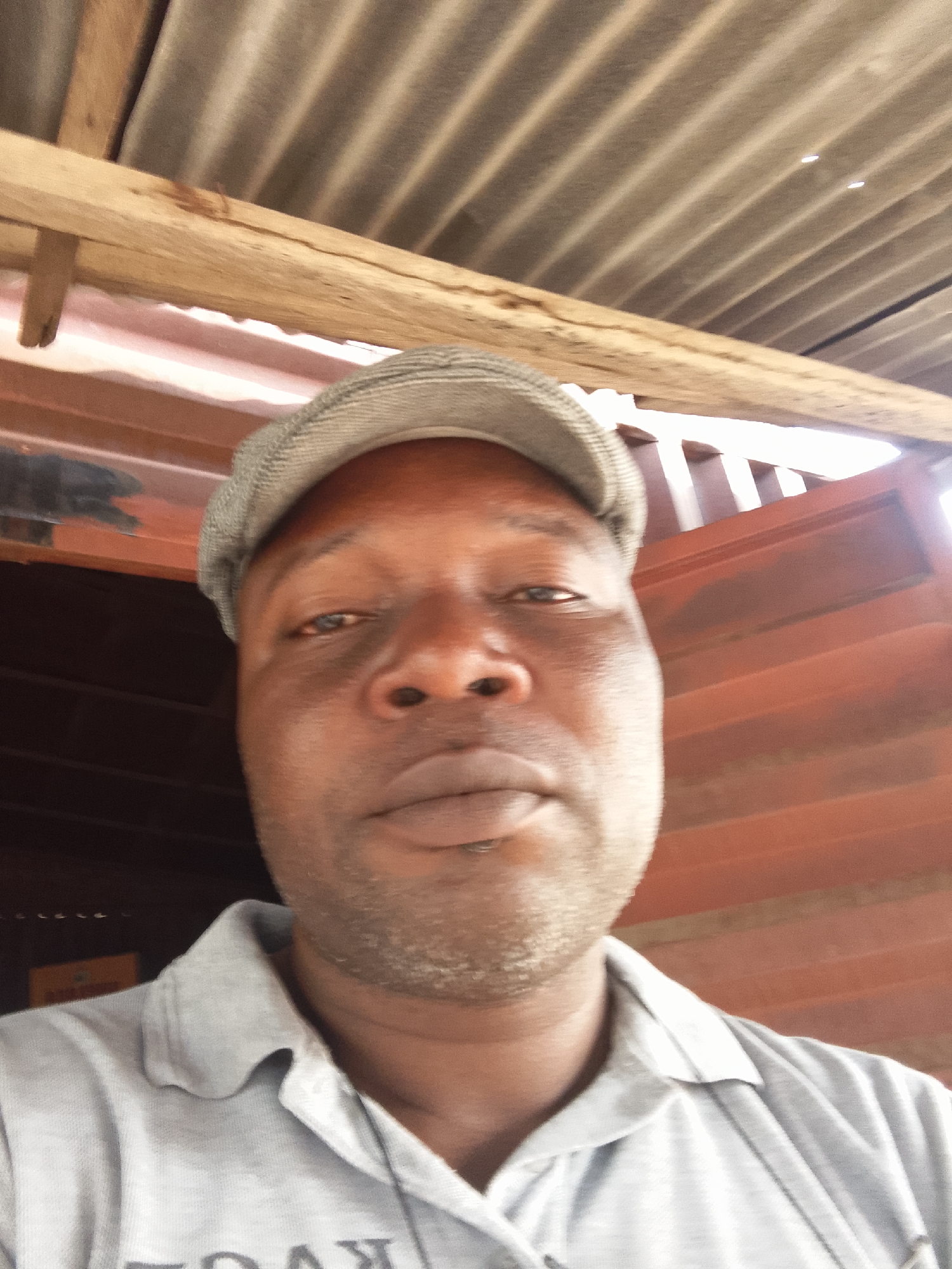 A close-up, low-angle portrait features a man with dark skin, wearing a light grey flat cap and a light grey polo shirt. He looks directly into the camera with a neutral expression. Bright light illuminates his face, particularly his forehead and nose, with some shadows visible under the brim of his cap.
The background primarily consists of the interior of a structure. Above the man's head is a corrugated metal ceiling with distinct ridges, supported by rough wooden beams. To the right and left sides of the frame, behind the man, are reddish-brown and dark brown structures or walls, possibly indicating metal panels or painted surfaces. Some light seems to be filtering in from the upper right and left, suggesting an open or translucent section in the structure.
Faint, reversed text is visible on the chest of the man's polo shirt, which appears to be "TOAK" or "YOAK". The image seems to be a selfie or a very close-up shot, focusing almost entirely on the man's face and the immediate overhead environment. The overall setting suggests a simple, functional covered space, possibly a workshop, market stall, or an unadorned room.