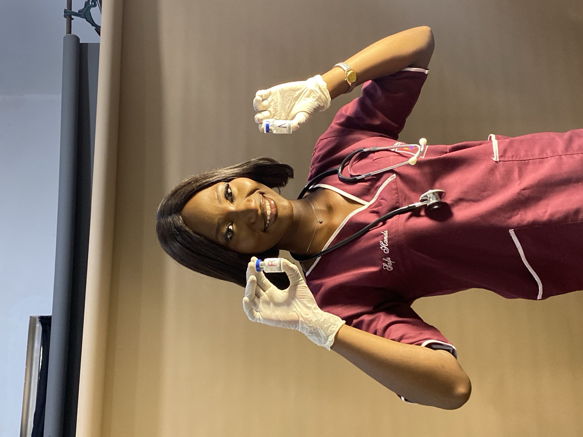 A young woman in Gura Top, Nigeria, wearing a maroon scrub suit with white trim and the words "Safe Hands" embroidered on the chest, is holding two vials in gloved hands. She is smiling and wearing a gold wristwatch. A stethoscope is around her neck. The background is a beige and grey studio backdrop.