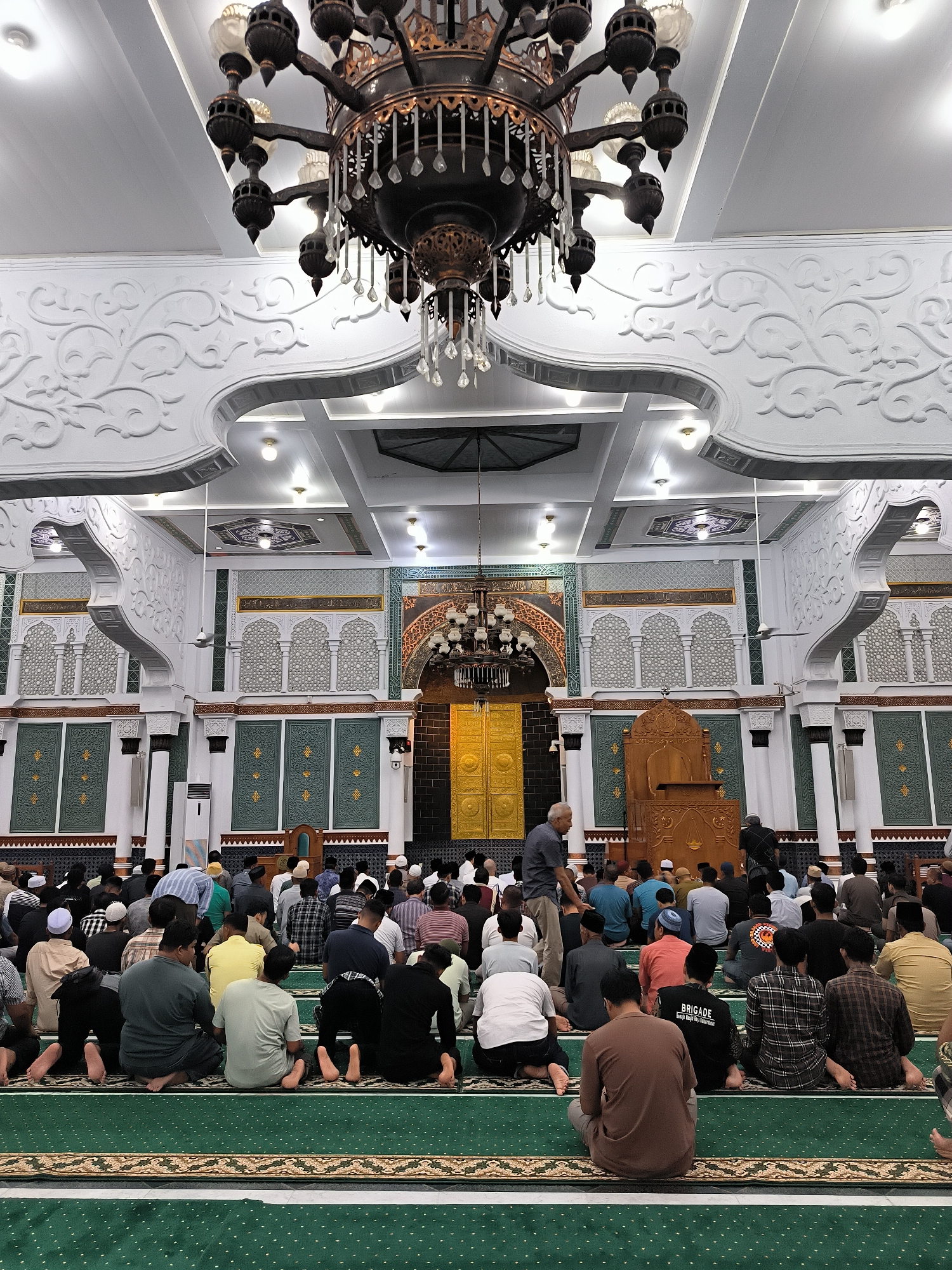 The image depicts the interior of a mosque, likely a place of worship in Banda Aceh, Indonesia, during prayer time. A large congregation of men is seated on a green carpet with intricate patterns, facing towards the front of the mosque where a golden-colored mihrab and a wooden mimbar are situated. The ceiling features ornate white stucco work and a large, dark chandelier. The walls are decorated with geometric patterns and calligraphy. The lighting suggests an indoor setting, possibly illuminated by overhead lights and natural light from unseen windows. The atmosphere appears solemn and communal, with individuals engaged in prayer or awaiting its commencement.