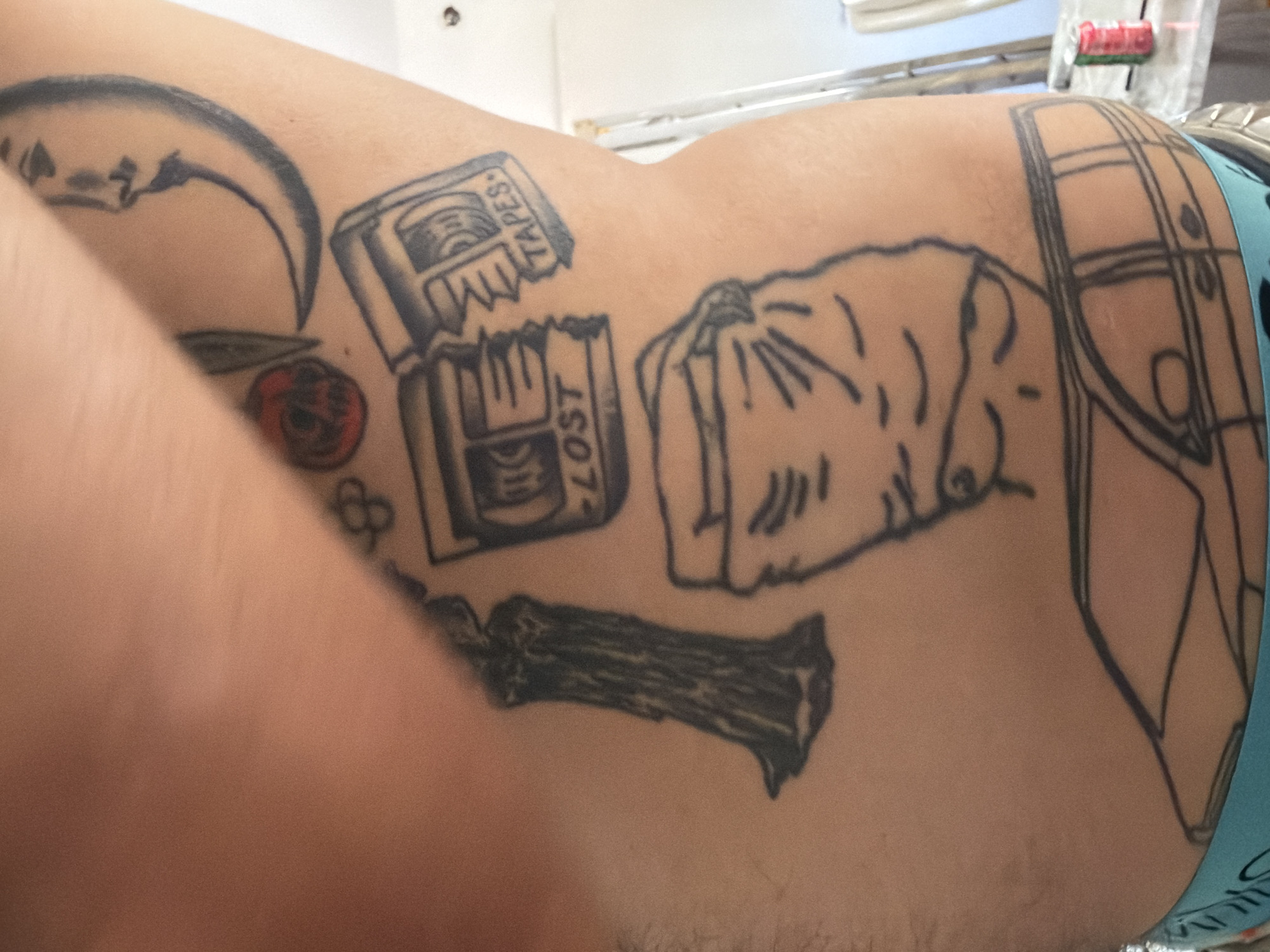 A full body shot of a person with numerous tattoos on their torso. The tattoos include a crescent moon with a face, a candle, two cassette tapes labeled "LOST" and "TAPES," a crumpled bag, and a car. The person is wearing blue underwear. The background is out of focus, but appears to be an indoor setting.