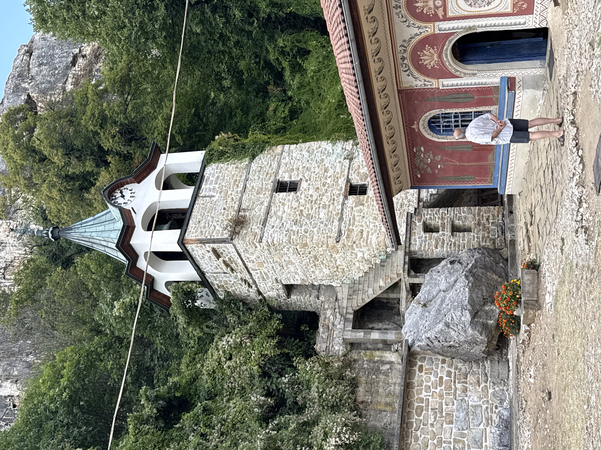 The image captures a scene at the Preobrazhenski Monastery, located near the village of Samovodene, Bulgaria. In the foreground, a man with graying hair, wearing a striped short-sleeved shirt and dark shorts, stands with his back to the camera, looking at a building with a red facade adorned with colorful murals and arched doorways. To the left, a stone bell tower rises, featuring a white belfry with arched openings and a teal-colored roof topped with a spire. Adjacent to the tower, a large boulder rests near a stone wall, with a set of stone stairs leading upwards. Lush green trees and vegetation surround the structures, and a rocky cliff face is visible in the background. The ground is paved with cobblestones and dirt.