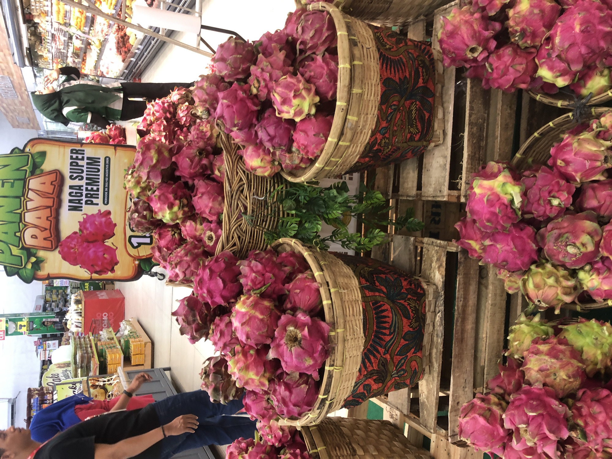 The image depicts a produce section of a supermarket in Langsa, Indonesia, specifically showcasing a large display of dragon fruit. In the foreground, three woven baskets overflow with vibrant pink dragon fruit, some of which have green accents. These baskets are placed on wooden crates. To the left, partially visible, is a man wearing a black t-shirt and blue jeans, accompanied by a woman in a blue headscarf and pink shirt. In the background, a large promotional sign for "Naga Super Premium" dragon fruit is prominently displayed, featuring an image of the fruit and text in Indonesian. A woman wearing a green hijab and a green uniform is visible near the produce display, seemingly interacting with the merchandise. Various shelves stocked with other food items and a ladder are also present in the background, suggesting a typical supermarket environment. The lighting suggests an indoor setting.