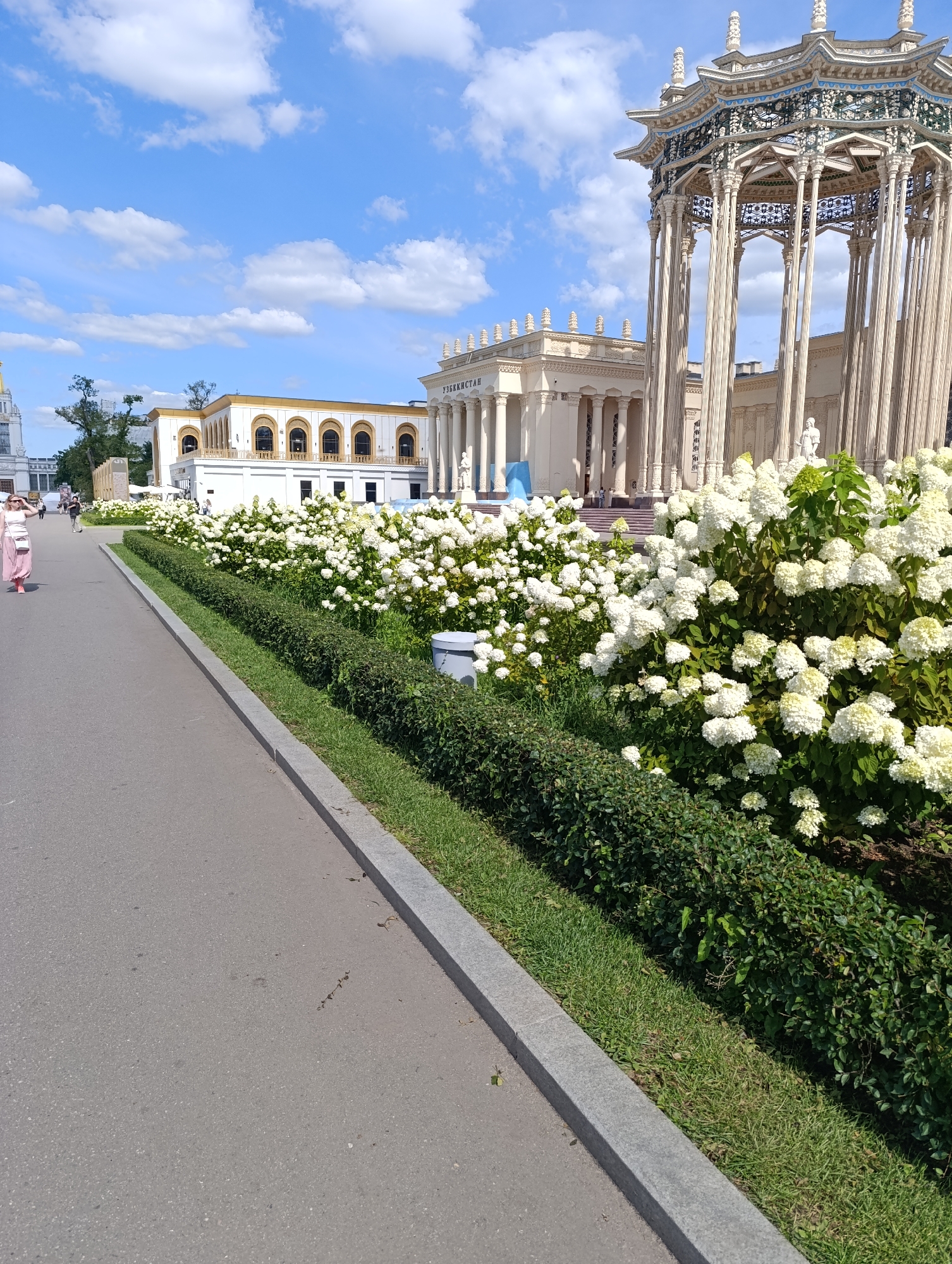 The image depicts a sunny day at an outdoor exhibition, likely in Moscow, Russia, as evidenced by the architecture characteristic of Soviet-era exhibition centers. In the foreground, a paved pathway runs alongside a meticulously maintained green hedge and a dense bed of white hydrangeas in full bloom. To the left, a woman in a pink skirt and white top walks away from the camera, her hand raised to shield her eyes.
In the background, a large, ornate pavilion with intricate latticework and numerous slender columns dominates the right side of the frame. Behind it, a classical building with a colonnade and arched windows stands, bearing the Cyrillic inscription "УЗБЕКИСТАН" (Uzbekistan), suggesting it is the Uzbekistan pavilion at the VDNKh (Exhibition of Achievements of National Economy) complex. The sky is a vibrant blue with scattered white clouds. Several other individuals are visible in the distance, walking along the pathway and near the buildings. The scene conveys a sense of a leisurely day at a public attraction.