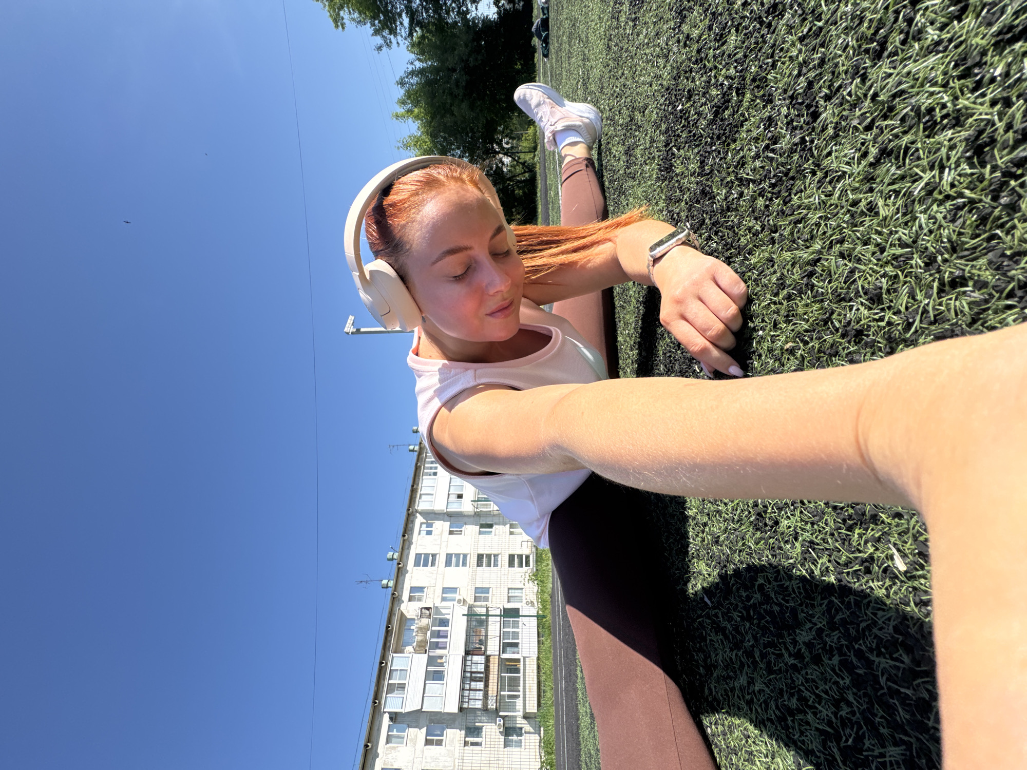 A young woman with red hair is performing a split on an artificial turf field. She is wearing a light pink tank top, brown leggings, and white headphones. She also has a smartwatch on her wrist. Behind her is a white apartment building and a clear blue sky. This photo was taken in the city of Chaikovsky, Russia.