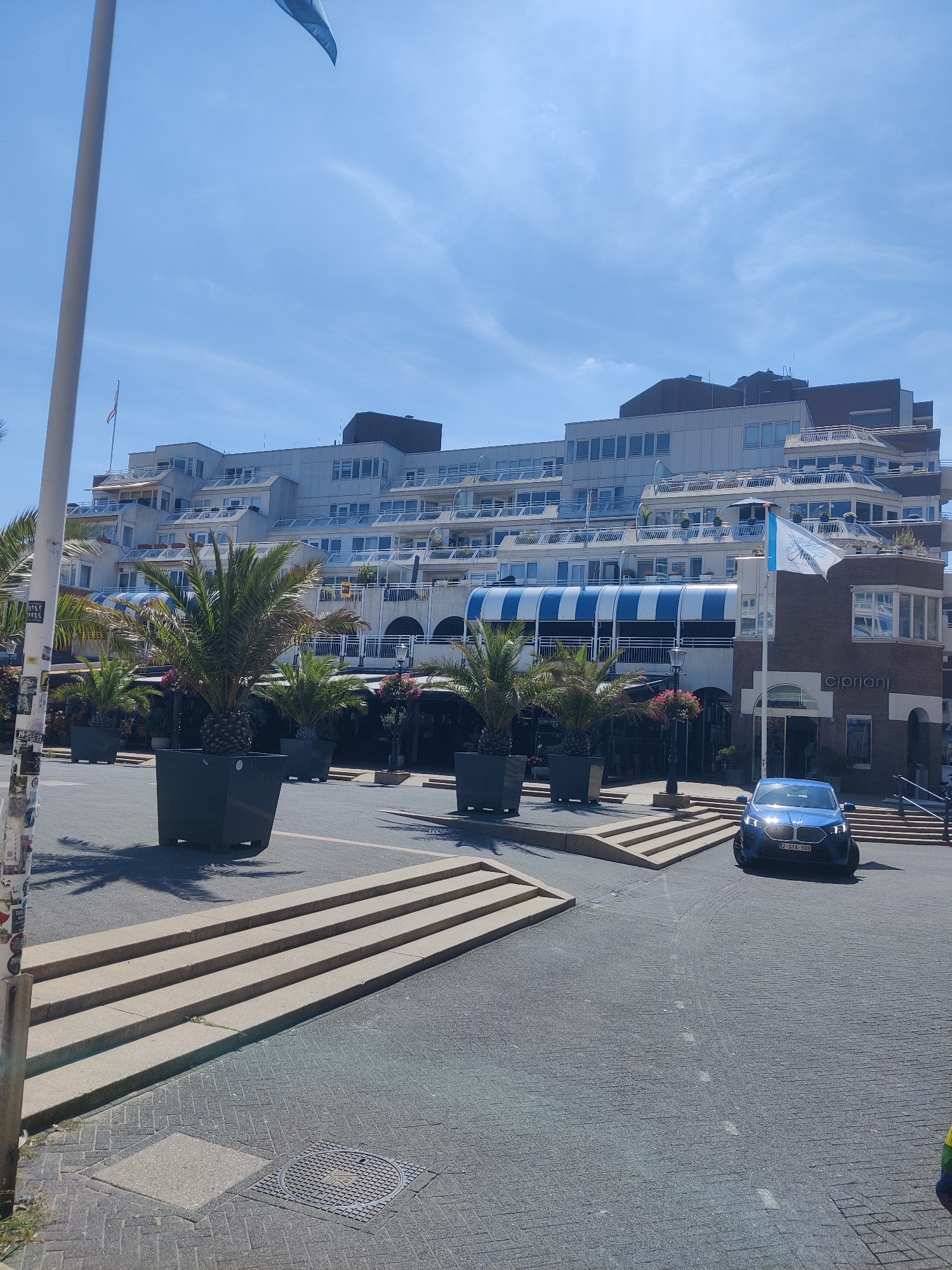 The image captures a view of the Cipriani restaurant, located on the waterfront in The Hague, Netherlands. A blue car is parked in front of the restaurant. The restaurant itself is a multi-story building with a distinctive facade featuring blue and white striped awnings. Palm trees in large planters are arranged in front of the restaurant, suggesting a seaside or resort location. The sky is clear blue with some scattered clouds, indicating a sunny day. The foreground shows a paved area with some concrete steps leading up to a higher level. The overall scene conveys a sense of a pleasant day out at a coastal establishment.