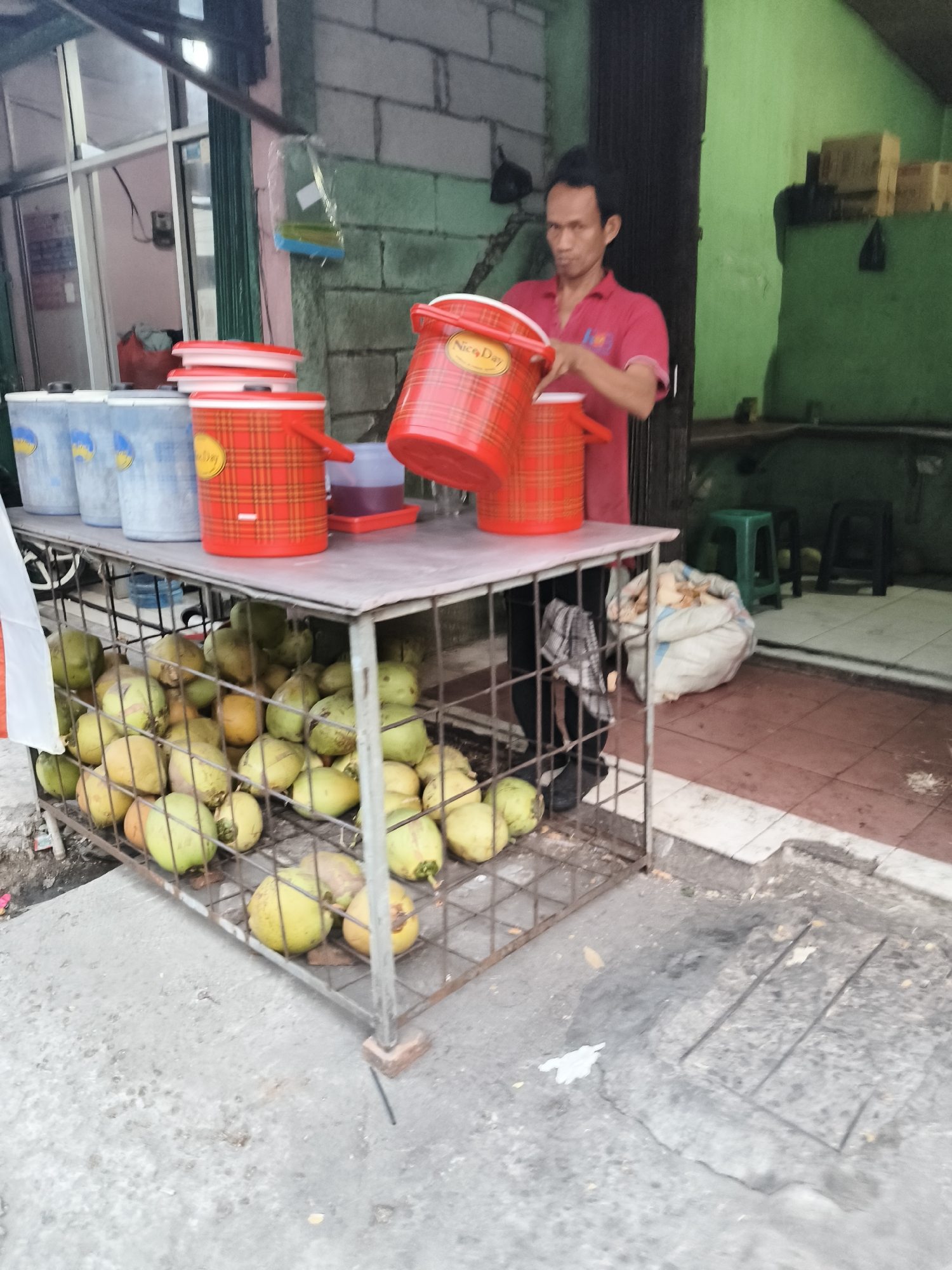 A street vendor stands behind a metal cage filled with coconuts, holding a red plastic container, in front of a small shop in South Tangerang, Indonesia. The vendor, a man wearing a red polo shirt, is in the process of pouring liquid from a red container into another one. The shop has a green wall and the floor is tiled with red and white. Several plastic containers are placed on a table. A white plastic bag is on the floor to the right of the vendor. The scene suggests a hot climate, likely during the day, as the coconuts are a refreshing drink.