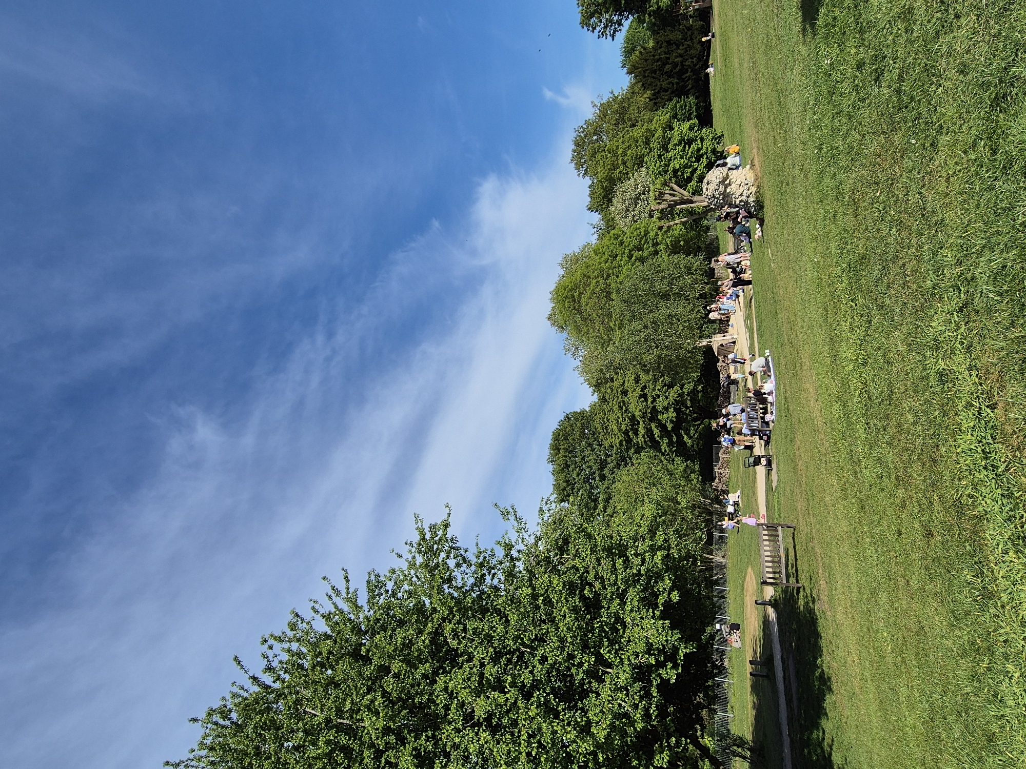 The image shows a scene in a park. The sky is blue with wispy white clouds. Lush green grass covers the right side of the image. Trees of various shades of green line the central area. People are congregated in the shade, some sitting on benches and others on the ground. A fence separates a path from the grassy area. The location is in West Suffolk, United Kingdom, based on geotagging information.