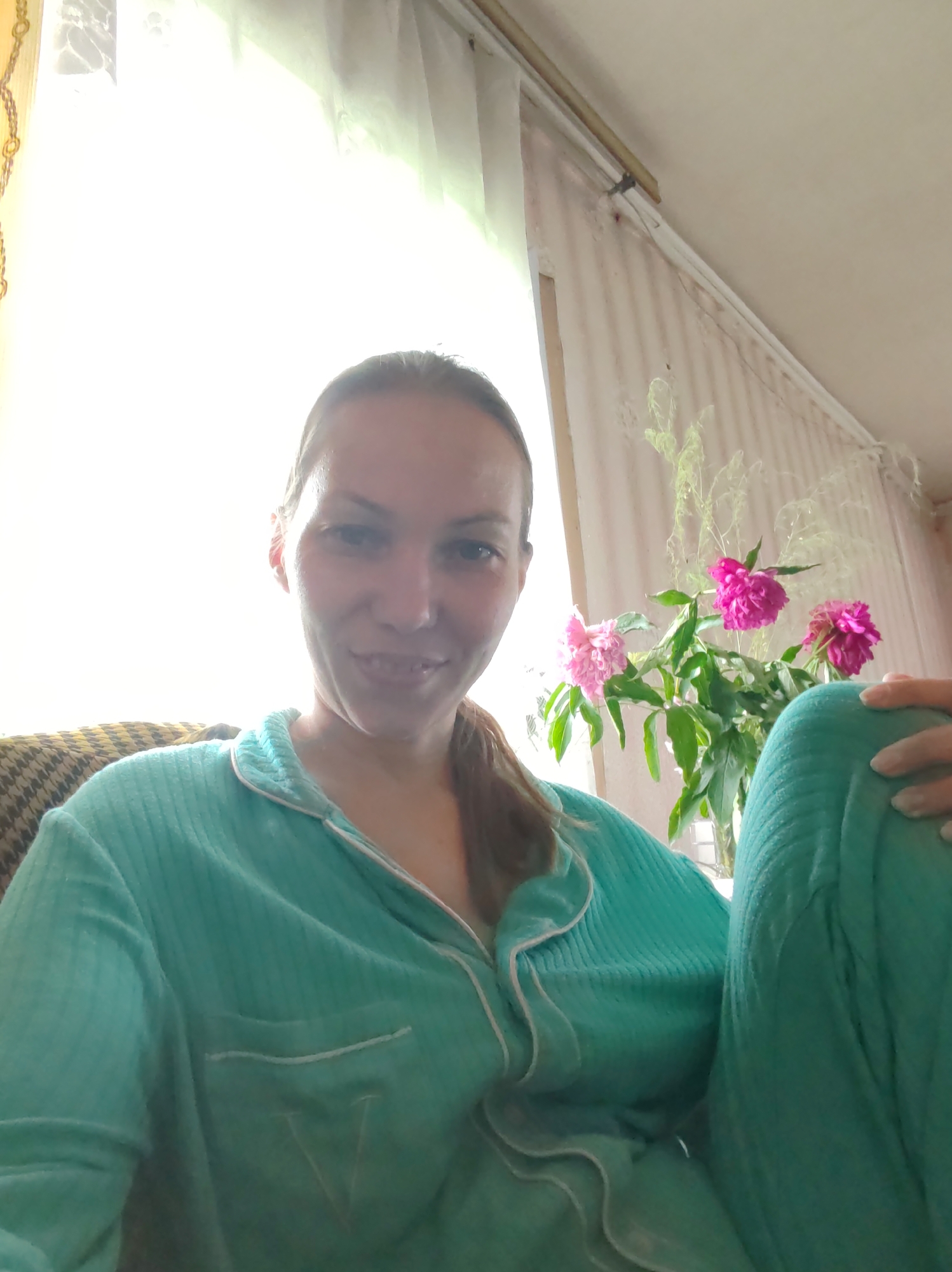 A selfie taken from a low angle shows a woman with long brown hair, wearing teal pajamas, smiling at the camera. She is seated on a couch with a brown and yellow patterned cushion behind her. To her right, on a table near a window with sheer curtains, are three pink peonies in a clear vase. The overall setting appears to be a domestic interior, possibly a living room, in the city of Chukhloma, Russia. The lighting suggests it is daytime, with light streaming in from the window.