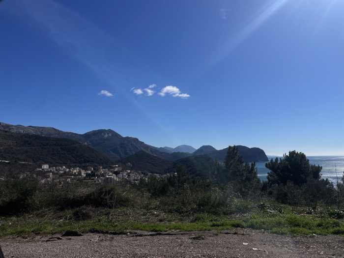 The image presents a panoramic landscape view from an elevated vantage point in Medinski Krš, Montenegro. The scene is captured under bright daylight conditions with a clear, deep blue sky, exhibiting a sun glare in the upper right quadrant.
The immediate foreground consists of rugged, uneven terrain composed of gray-brown dirt and small rocks, sparsely covered with patches of light green grass and low-lying, dark green scrub vegetation.
The midground reveals a coastal settlement and geographical features. To the left, a densely packed cluster of multi-story buildings, predominantly with light-colored facades, forms a town situated against the lower slopes of mountains. To the right, several tall, dark green coniferous trees obstruct a portion of the view toward the sea. Beyond these trees, a vast expanse of calm, turquoise-blue water, identified as the sea, reflects sunlight. A prominent, dark green, rugged peninsula or island extends into the sea from the right side of the frame.
The background is characterized by a range of dark, heavily forested mountains that rise behind the town and stretch across the horizon. The sky above is uniformly clear blue, with only two small, distinct white cumulus clouds visible above the central mountain peaks. No human figures, animals, or active subjects are present in the image.