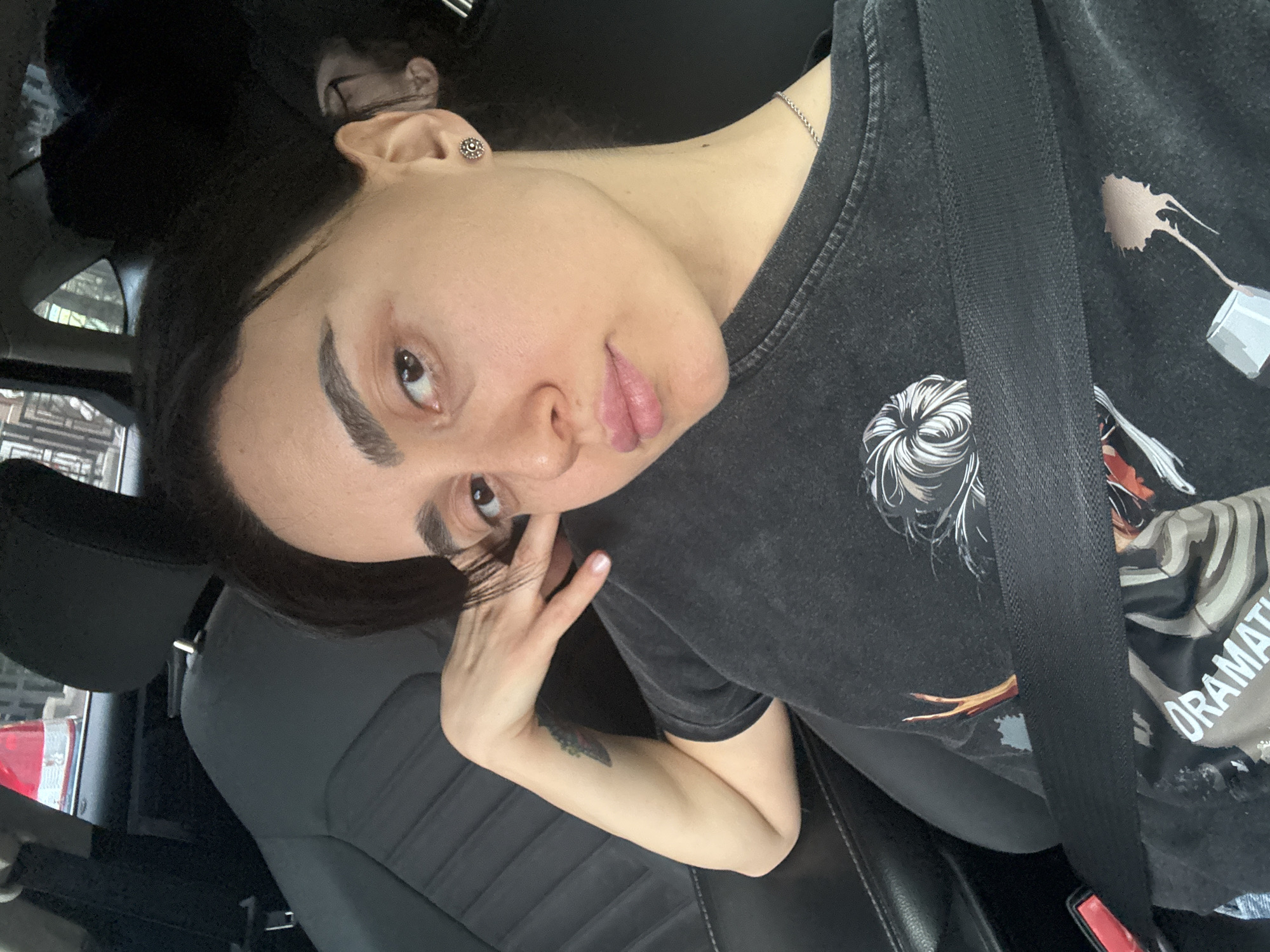 A young woman is inside a car, presumably in Tbilisi, Georgia. The city is alluded to by the exterior view from the car windows. She wears a dark-colored T-shirt, and a seatbelt is fastened across her chest. She is looking at the camera, with her left hand touching her face near her eye. Her hair is dark and pulled back. She has a small stud earring in her left ear and a delicate silver chain necklace. The car's interior features dark leather seats.