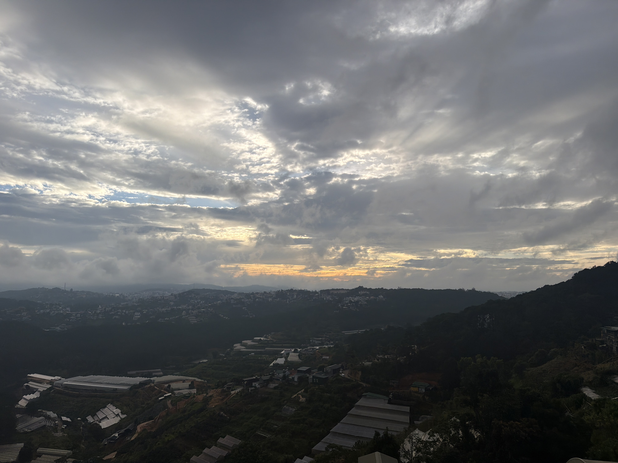 The image presents a panoramic view of a mountainous city at dusk, likely Xuân Trường, Đà Lạt, Vietnam. The sky is dominated by dramatic, overcast clouds, with hints of a muted orange and yellow sunset peeking through the gaps, creating a diffused light across the landscape. Below the sky, a sprawling city unfolds, nestled within the dark, rolling hills. Buildings, densely packed in the distance, are visible as a cluster of lights and structures. Closer to the foreground, the terrain slopes steeply, revealing agricultural greenhouses with white, translucent roofs, indicative of local farming practices. The lush, dark green foliage of trees and vegetation covers the hillsides, interspersed with more scattered buildings and structures. The overall impression is one of a vast, natural landscape transitioning into human settlement under a moody, atmospheric sky.