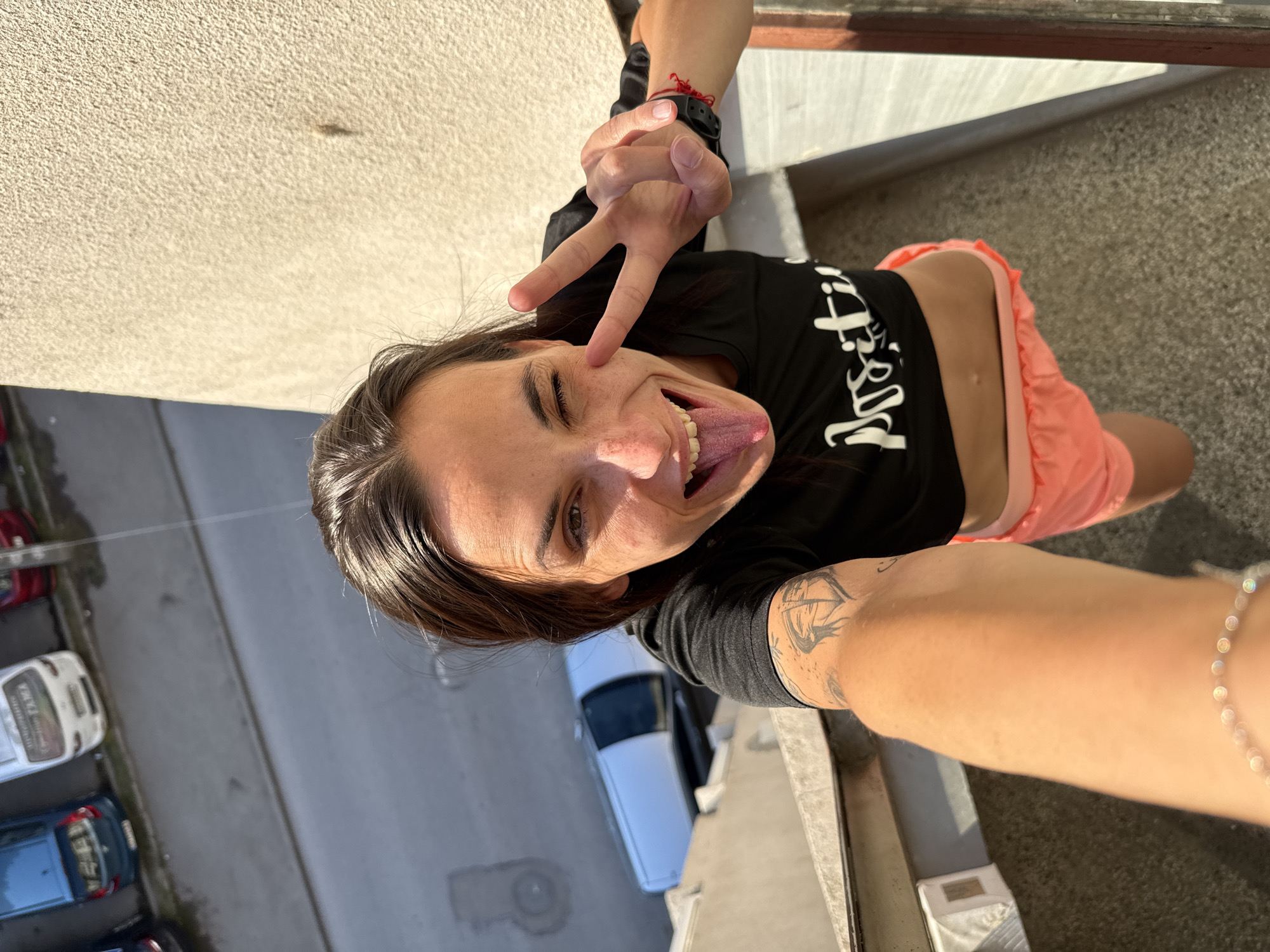 A person is taking a selfie from above, sticking out their tongue and making a peace sign. The person has dark hair and is wearing a black shirt and pink shorts. They are standing on a balcony or ledge, looking down at a street with cars parked on it. The location is Pleven, Bulgaria.