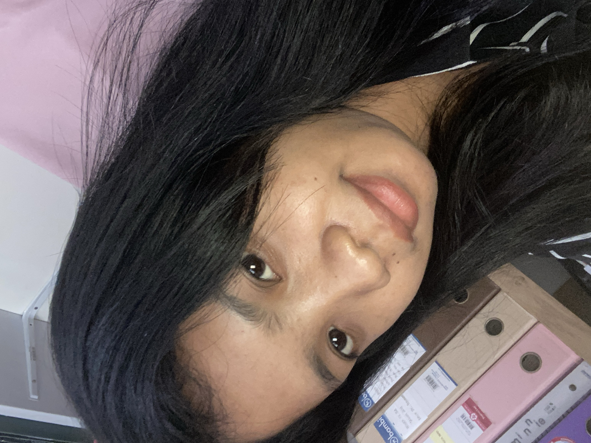 A person with dark hair is posing for a selfie. The background includes a pink surface and a collection of folders. The location is potentially Negara, Indonesia, given the Bambi brand on a folder.