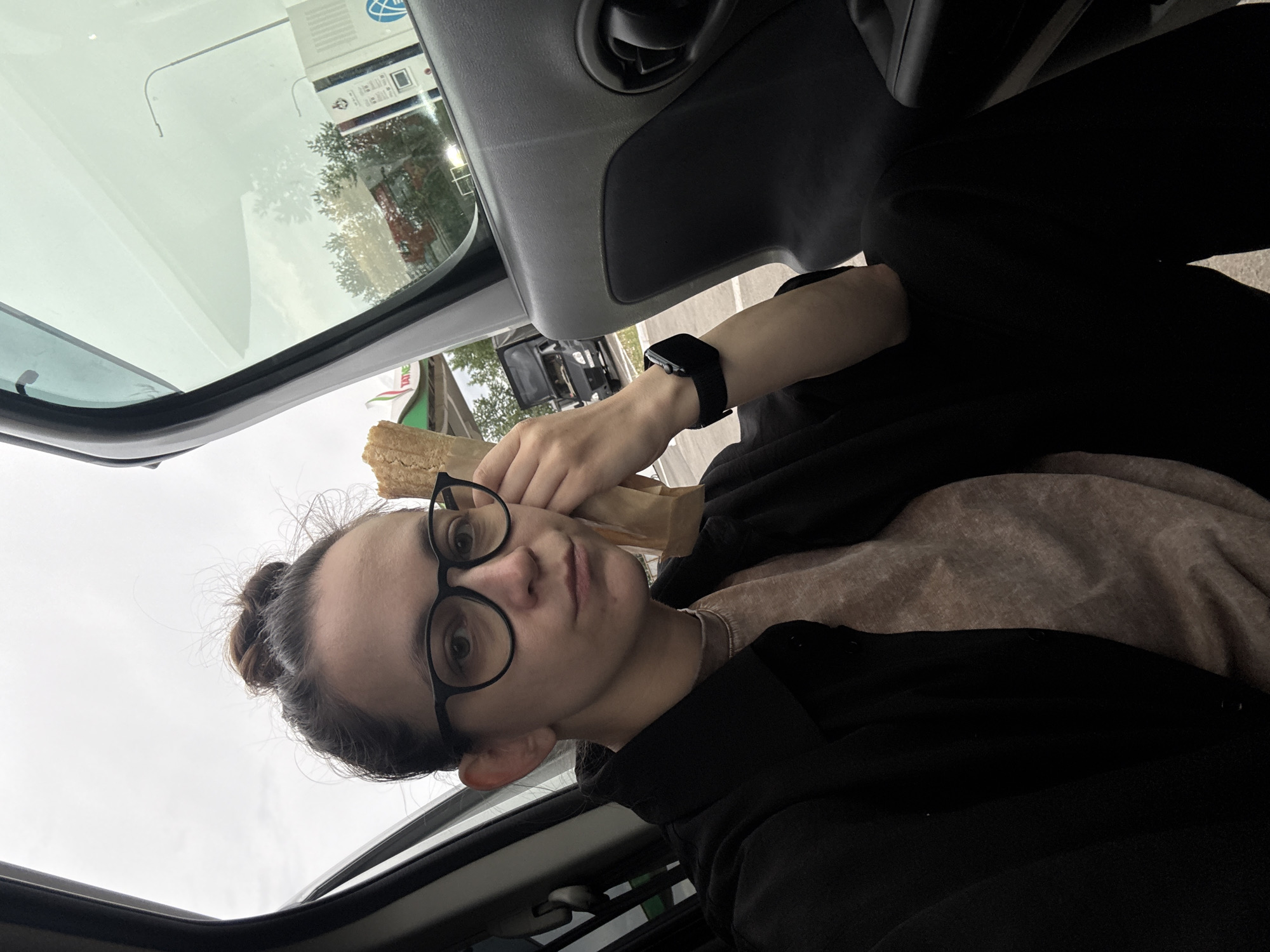 A person with glasses, a bun hairstyle, and a watch on their left wrist is sitting inside a vehicle. They are holding a wrapped food item. The environment appears to be a gas station, as indicated by the gas pump visible through the open sunroof. The location is identified as the city of Unknown, Russia.