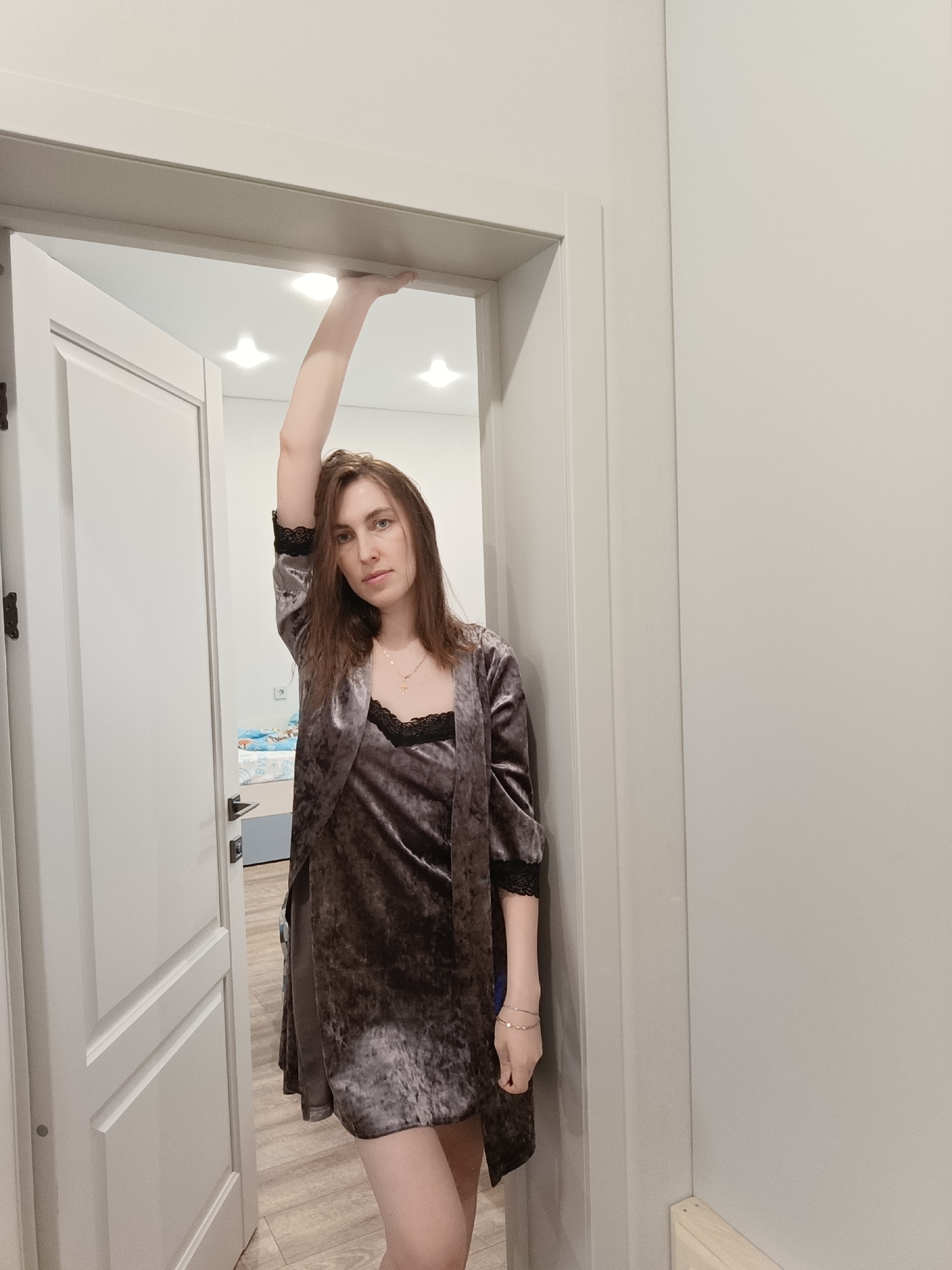 A young woman stands in a doorway. She is wearing a velvet dress and robe with black lace trim. Her right arm is raised, her hand touching the doorframe. She has long brown hair and is wearing a necklace and bracelets. The doorway leads into a room with a bed visible in the background. The walls are painted a light color, and there are light fixtures on the ceiling. The location is implied to be in a home in Chaikovsky, Russia.