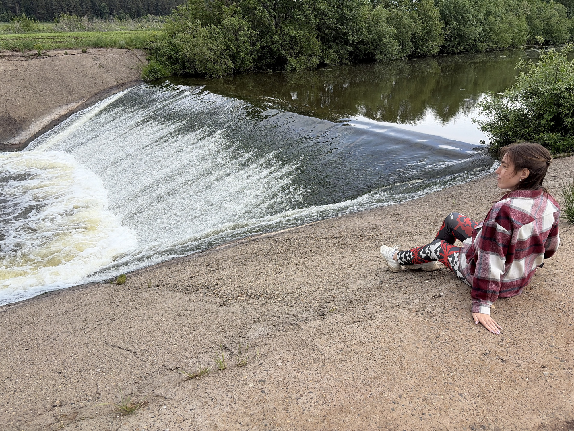 A young woman sits on a concrete dam wall in Nizhnii Vishur, Russia. The water from the dam cascades down in a wide sheet of white foam. The woman is wearing patterned leggings, white sneakers, and a red and white flannel shirt. She is looking out towards the water and the green foliage on the other side of the dam. The background includes trees and a river. The ground is a light greyish-brown concrete.