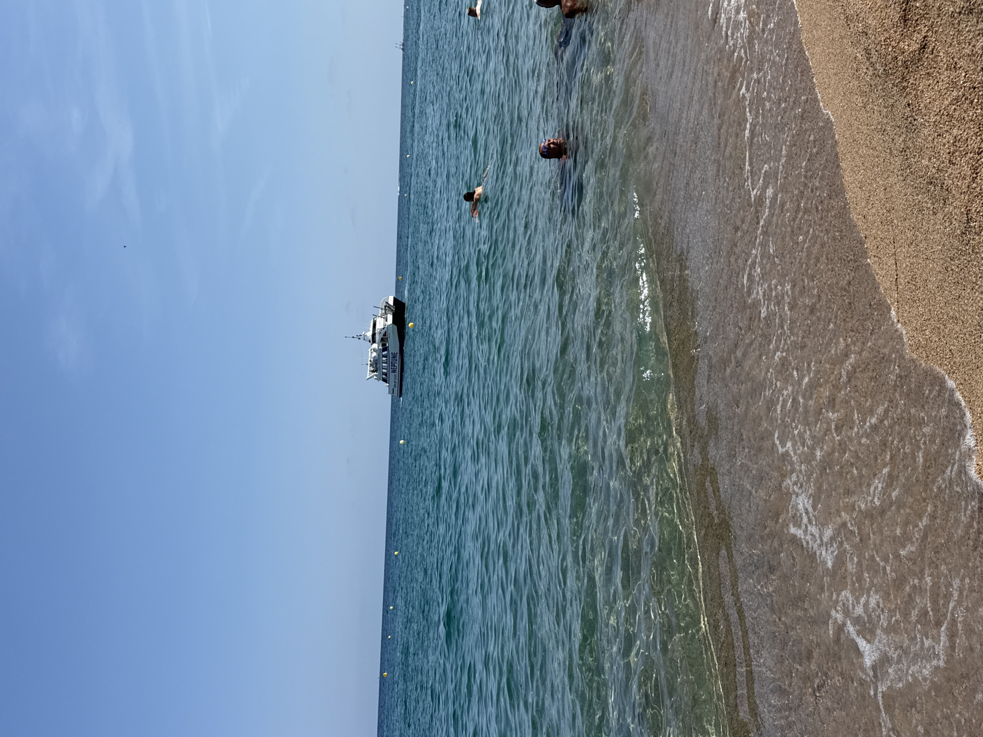 The image depicts a sunny day at a beach in Blanes, Spain. The foreground shows the sandy shore with gentle waves lapping at the edge. In the middle ground, several people are swimming and enjoying the clear, turquoise water. A white boat with "NEPTUNO" written on its side is visible in the background, further out at sea, along with a few yellow buoys marking the swimming area. The sky is a clear, vibrant blue with a few faint wisps of clouds. The overall scene is one of a peaceful and pleasant day at the coast.