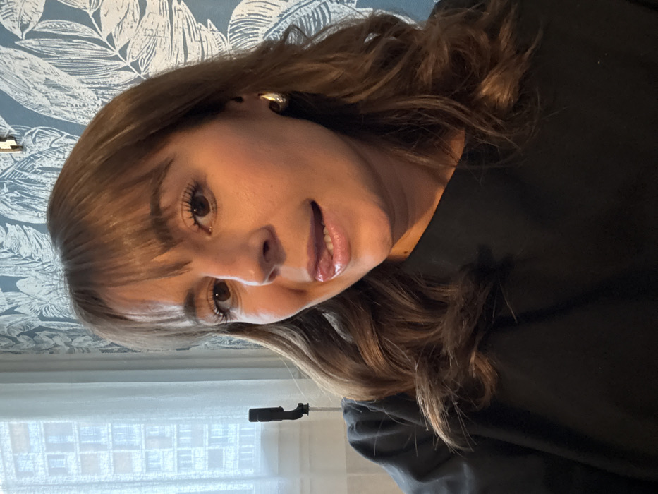 A woman with brown hair and bangs looks at the camera. She wears a black top and golden hoop earrings. Her lips are slightly parted, revealing a glimpse of her teeth. The background includes a white curtain, a window with a building view, and a wall with a blue and white leaf pattern. The image is rotated slightly.