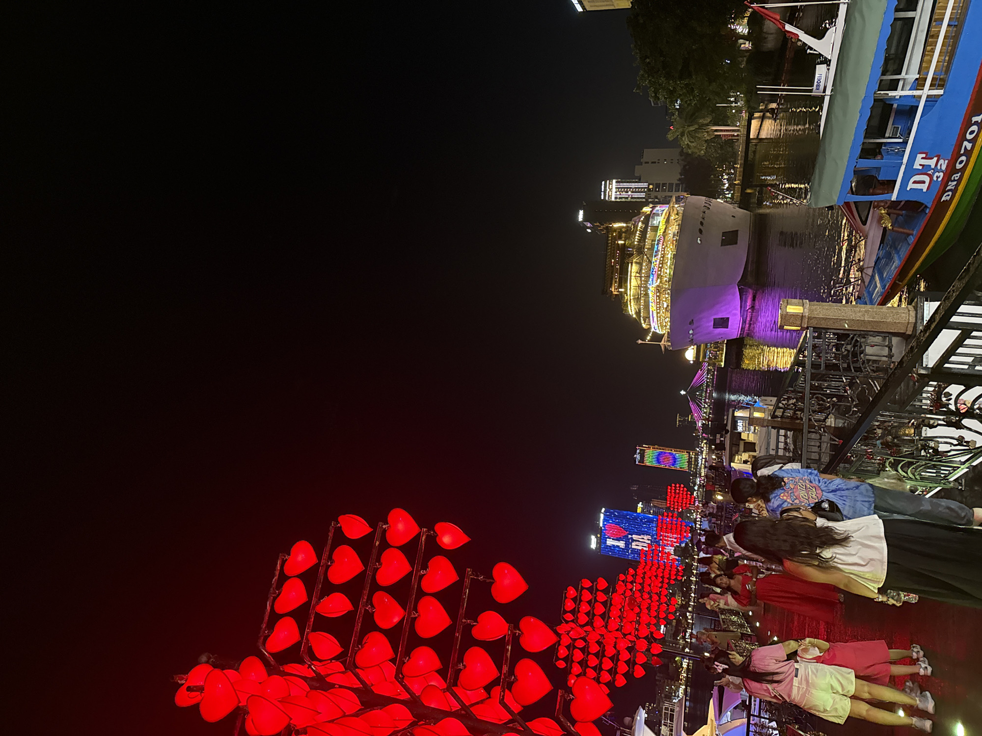 Nighttime scene in Đà Nẵng, Vietnam. Red heart-shaped lanterns illuminate the foreground. People are walking. Buildings with lighted displays are visible in the background. Boats are docked on a river; the main boat has colorful lights. The sky is dark.