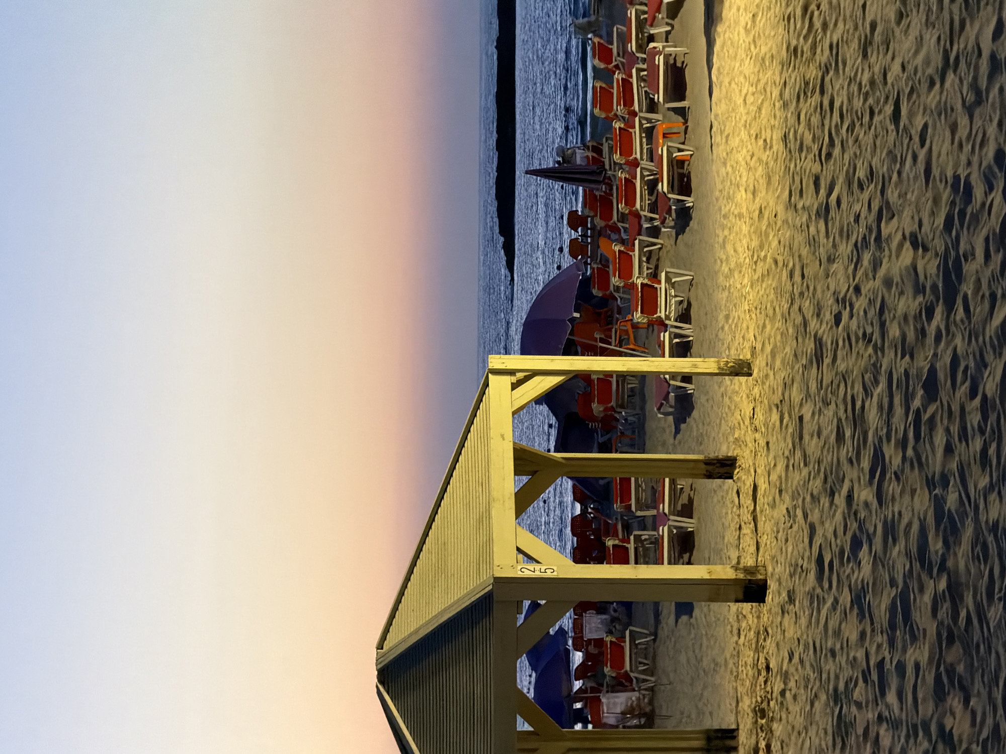 The image captures a beach scene in Tel-Aviv, Israel, likely at dusk or dawn based on the gradient sky from pale blue to orange-pink. In the foreground, the corner of a wooden beach hut or shelter is visible with the number "25" painted on it. Adjacent to the structure are rows of red and white beach chairs, some with closed purple umbrellas. The chairs appear unoccupied and are arranged neatly in rows on the sand. The sand itself is illuminated, showing pronounced textures and patterns, and extends into a dark area. Beyond the beach chairs and umbrellas, the sea forms the background. The water is relatively calm with faint ripples.