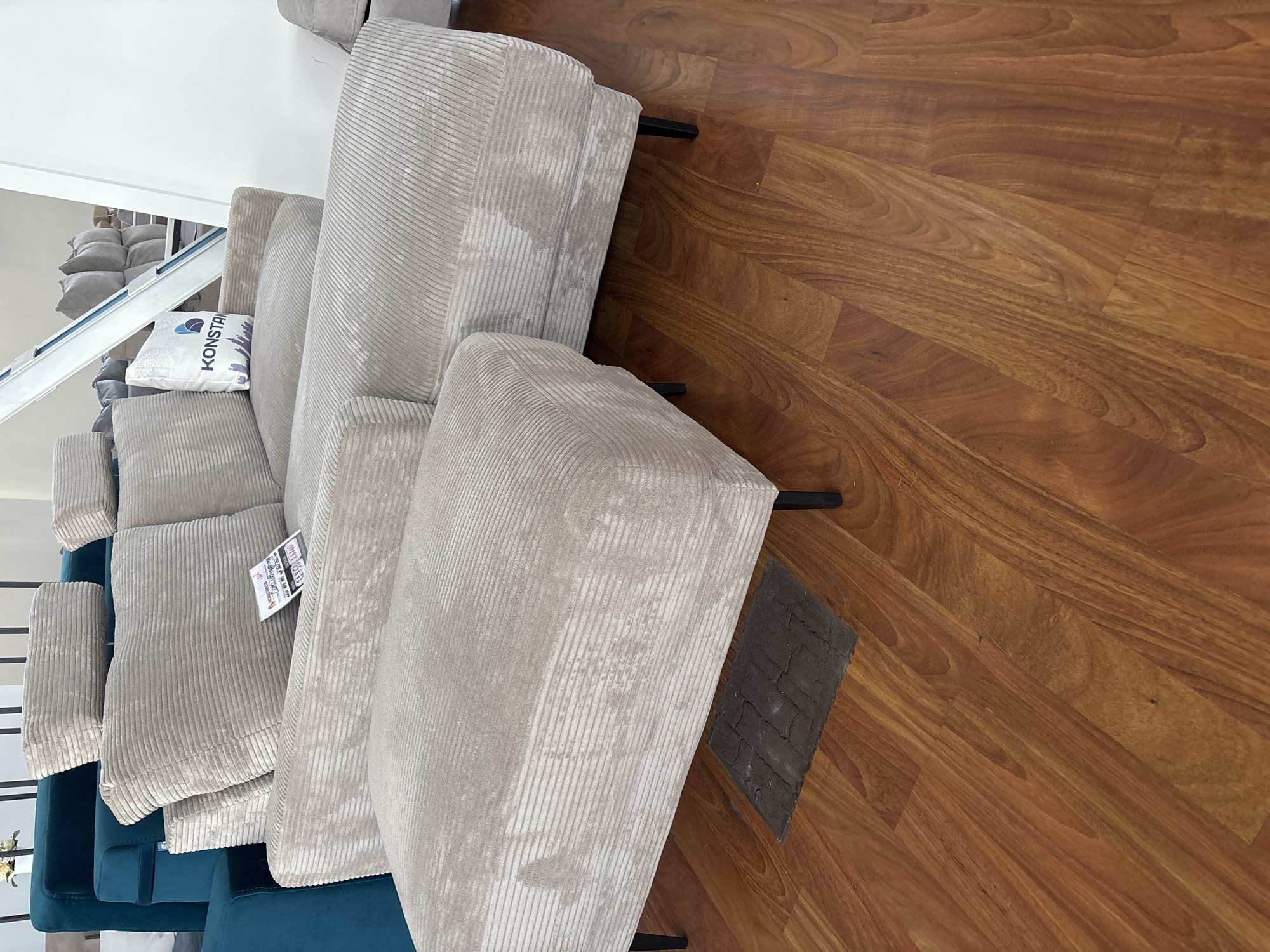 The image shows a close-up view of several plush, beige corduroy sofa sections placed on a polished wooden floor. The sofa sections are arranged in a staggered manner, with some overlapping others. One of the sofa sections has a white tag attached to it with blue and purple text and a graphic of buildings. Another tag, this one orange and white with black text, is visible on a different sofa section. A small, dark gray, textured object, possibly a mat or a piece of flooring, is positioned on the wooden floor between two of the sofa sections. The wooden floor exhibits a warm, reddish-brown hue with a distinct grain pattern. The lighting suggests an indoor setting, likely a showroom or a well-lit room. The perspective is from above and to the side, looking down at the sofa and floor. The overall impression is one of a clean, modern interior space, possibly a furniture store or a home. There are no people visible in the image. The scene is static and does not depict any activity. The location is specified as Zaporizhzhia, Ukraine.