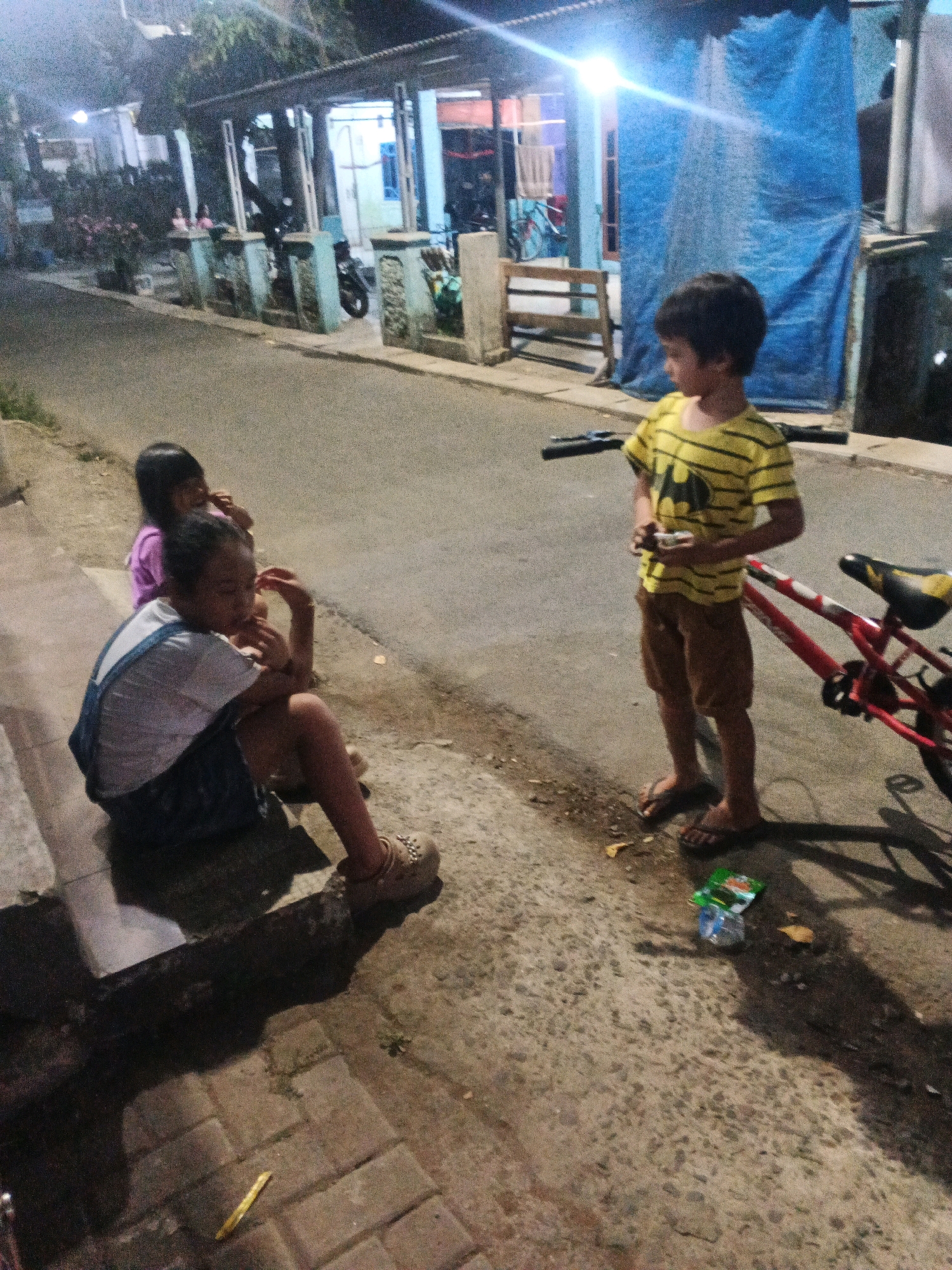 The image depicts an outdoor scene at dusk, possibly in Kadu, Indonesia. In the foreground, two young girls are sitting on a raised concrete ledge beside a paved road. The girl on the left is wearing denim overalls over a white shirt and appears to be resting her head on her hand. The girl on the right is wearing a purple shirt. Behind them, a young boy wearing a yellow t-shirt with a black Batman logo and brown shorts stands near a red bicycle. He is holding a small object in his hands. The environment includes a road, sidewalk, and buildings with a blue tarp draped over part of one structure. There are shadows cast on the ground, indicating the low angle of the light. Scattered debris, including a plastic bottle and a packet, is visible on the ground near the boy and the bicycle.