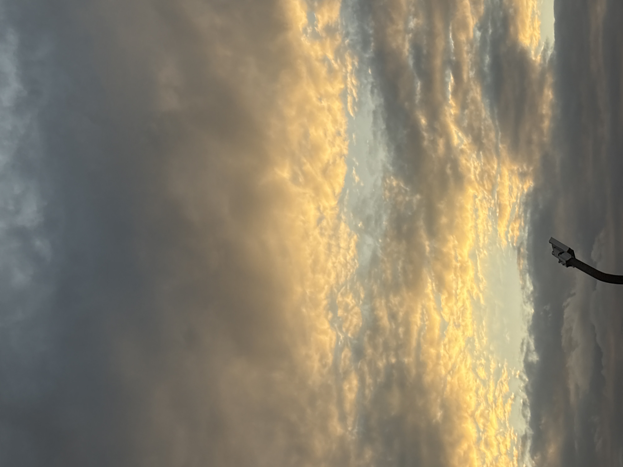The image captures a dramatic sky over Georgiyevsk, Russia, characterized by a mix of dark, brooding clouds and patches of warm, golden light, suggesting either sunrise or sunset. In the foreground, a portion of a dark, curved pole is visible, topped with what appears to be a detached streetlamp fixture. The fixture is white with metallic accents, and its shape is angular. The rest of the image is dominated by the expansive sky filled with textured clouds, ranging in color from deep grey to vibrant yellow and orange, with hints of blue peeking through in some areas. No people or animals are visible.