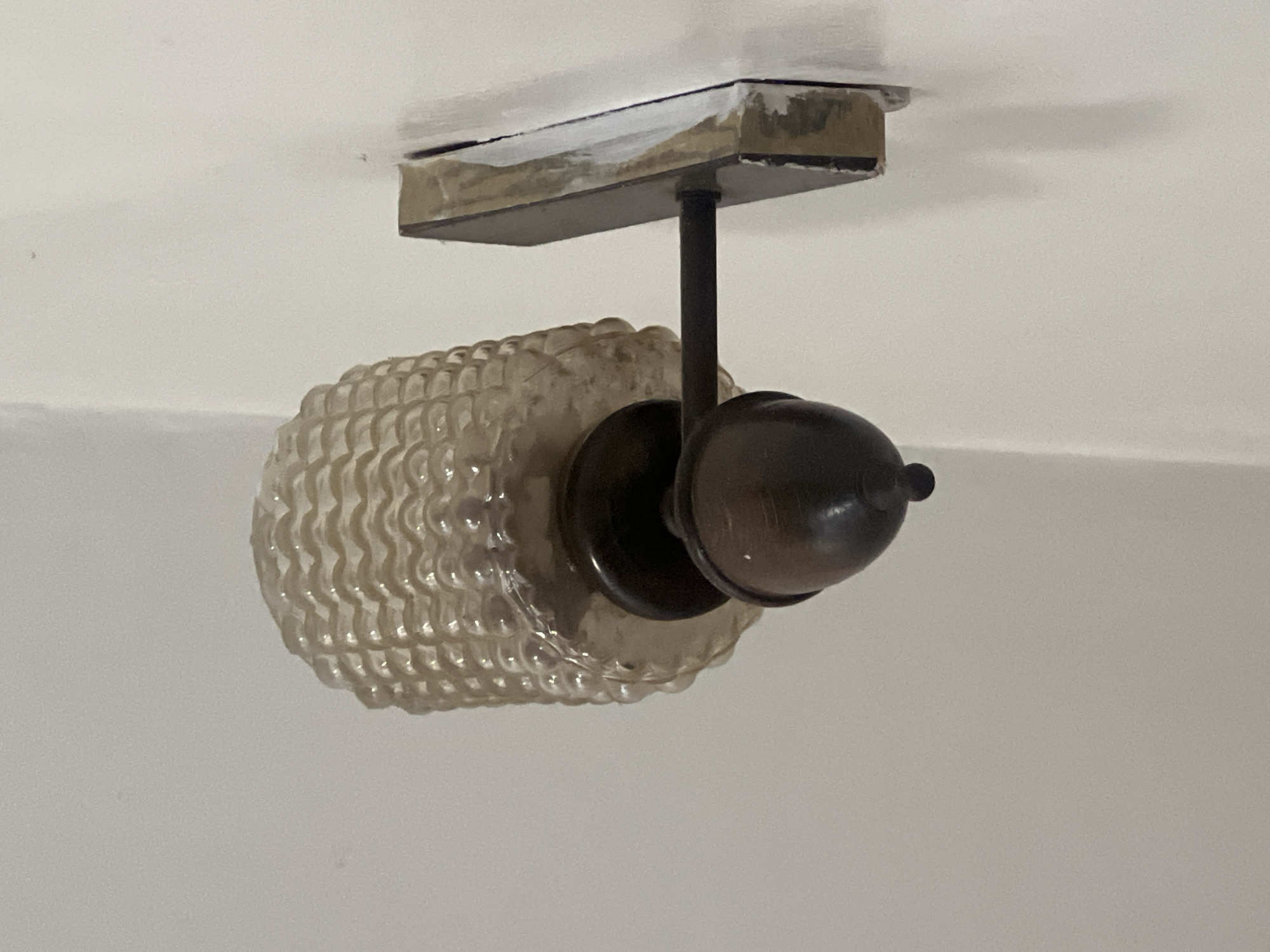 This image depicts a wall-mounted light fixture, known as a sconce, installed on an interior white wall in Casablanca, Morocco. The fixture features a translucent glass shade with a wavy, textured, or corrugated pattern, appearing a light amber or clear hue and showing signs of age or dust accumulation. Below the shade, a dark-colored, possibly bronze or dark brown, metal support arm extends horizontally from a rectangular backplate. A decorative, bell-shaped finial or housing, also dark-colored, is positioned beneath the light source, culminating in a small, spherical detail at its lowest point. The rectangular backplate is affixed to the wall, but the installation reveals visible gaps and white residue, consistent with dried caulk, plaster, or paint, around its edges where it meets the white wall surface. No individuals or active elements are present in the scene, which is illuminated by ambient light.