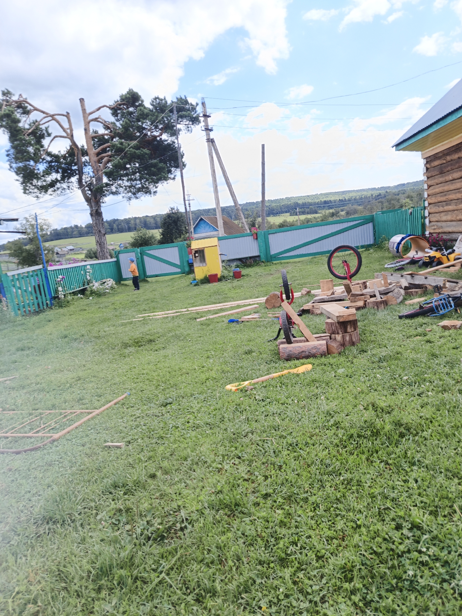 The image depicts an outdoor scene in Islambaevo, Russia, likely a rural or semi-rural setting. A wooden log house with a green-trimmed roof is partially visible on the right side of the frame. In the foreground and midground, a grassy yard is covered with various construction materials, including lumber, logs, and dismantled wooden structures. Several tools are also scattered across the grass, such as a yellow shovel, a leaf blower, and parts of what appear to be a bicycle or unicycle. A child wearing an orange shirt and a blue cap stands near a green fence in the middle ground, looking towards the left. Beyond the fence, a tree with distinctive, spreading branches stands tall, and power lines run across the sky. The background shows rolling hills and a hazy, rural landscape. The sky is partly cloudy with patches of blue.