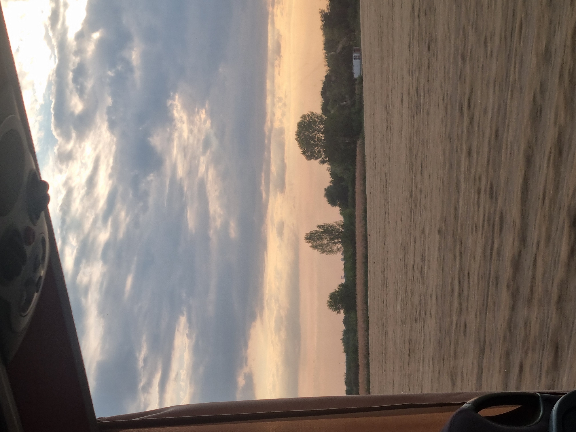 The image shows a view from inside a vehicle, likely a bus, looking out at a rural landscape. The sky is filled with dramatic clouds in shades of gray and white, with patches of sunlight breaking through, indicating either sunrise or sunset. The horizon is lined with trees and foliage, with a field of dry grass or crops in the foreground. A small white building is visible among the trees on the right side of the image. The overall impression is one of a serene, natural environment. The provided location "Mădăras, Romania" suggests this scene is situated in that region.
