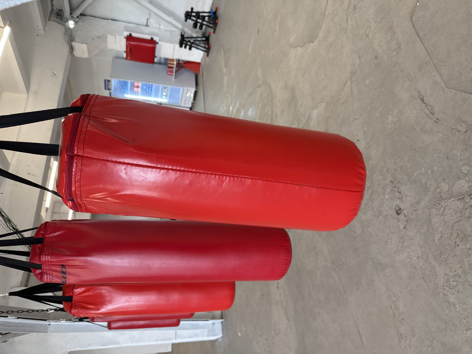 The image depicts a series of red punching bags hanging in what appears to be a gym or training facility. The bags are cylindrical and suspended by black straps. In the background, partially out of focus, there is a vending machine, workout equipment, and the architecture suggests the location might be in an industrial or commercial building. The scene is in Sin'kovo, Russia. The environment is utilitarian, and the lighting is bright. There are no people visible in the frame.