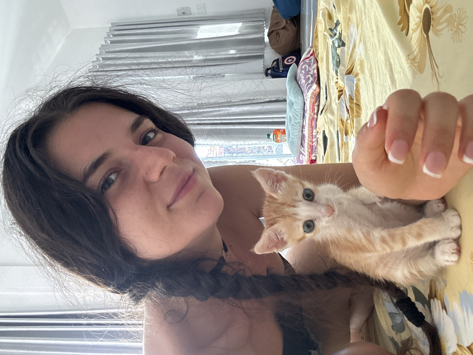 A fair-skinned woman with dark, long hair styled in a braid is holding an orange tabby kitten in her lap. The kitten, with blue eyes, faces the camera. The woman is wearing a dark-colored top. A floral-patterned yellow blanket or sheet serves as the backdrop. In the background, there is a window with blinds, luggage, and other items. The location could be Mũi Né, Vietnam, although there are no defining landmarks visible to confirm the location. The interaction between the woman and the kitten suggests affection or care.