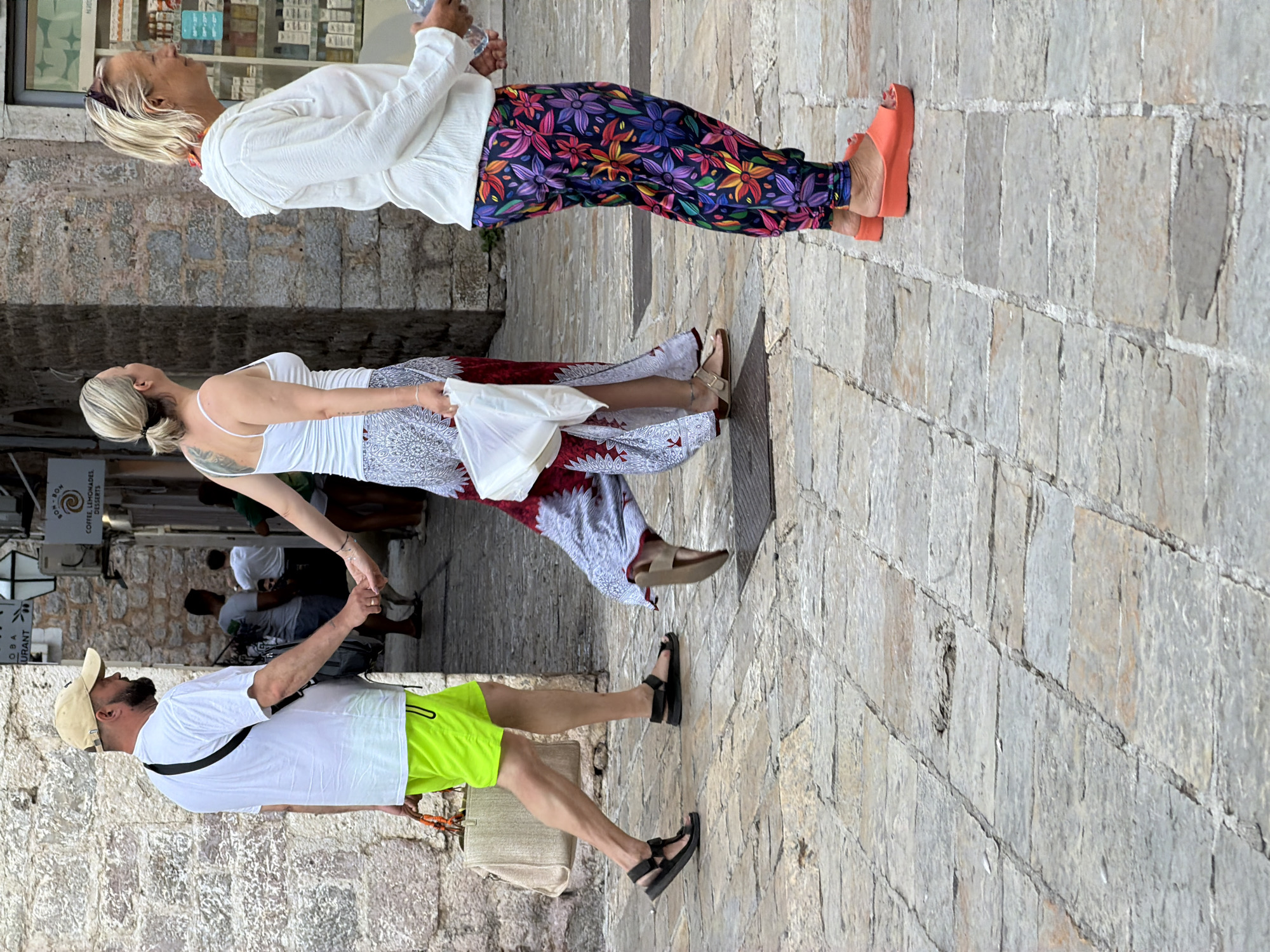 This is a street-level, eye-level, medium shot of three people walking on a cobblestone street in Budva, Montenegro. The people are walking towards the right of the frame, away from the viewer. In the foreground, a man wearing a white t-shirt, a beige cap, and bright neon green shorts walks with a beige bag slung over his left shoulder and holding a bag in his right hand. He is wearing black sandals. Beside him, a woman in a white tank top and a long, patterned skirt walks hand-in-hand with the man, carrying a white plastic bag. She is wearing brown sandals. To the right of these two individuals, a woman in a white hooded jacket and brightly colored floral pants walks with a water bottle in her right hand. She is wearing orange platform sandals. The background shows a stone building with a storefront, possibly a cafe, as suggested by signage reading "COFFEE, LEMONADES, DESSERTS." Other people are visible in the background, suggesting a busy urban environment. The sunlight casts shadows on the cobblestone street.