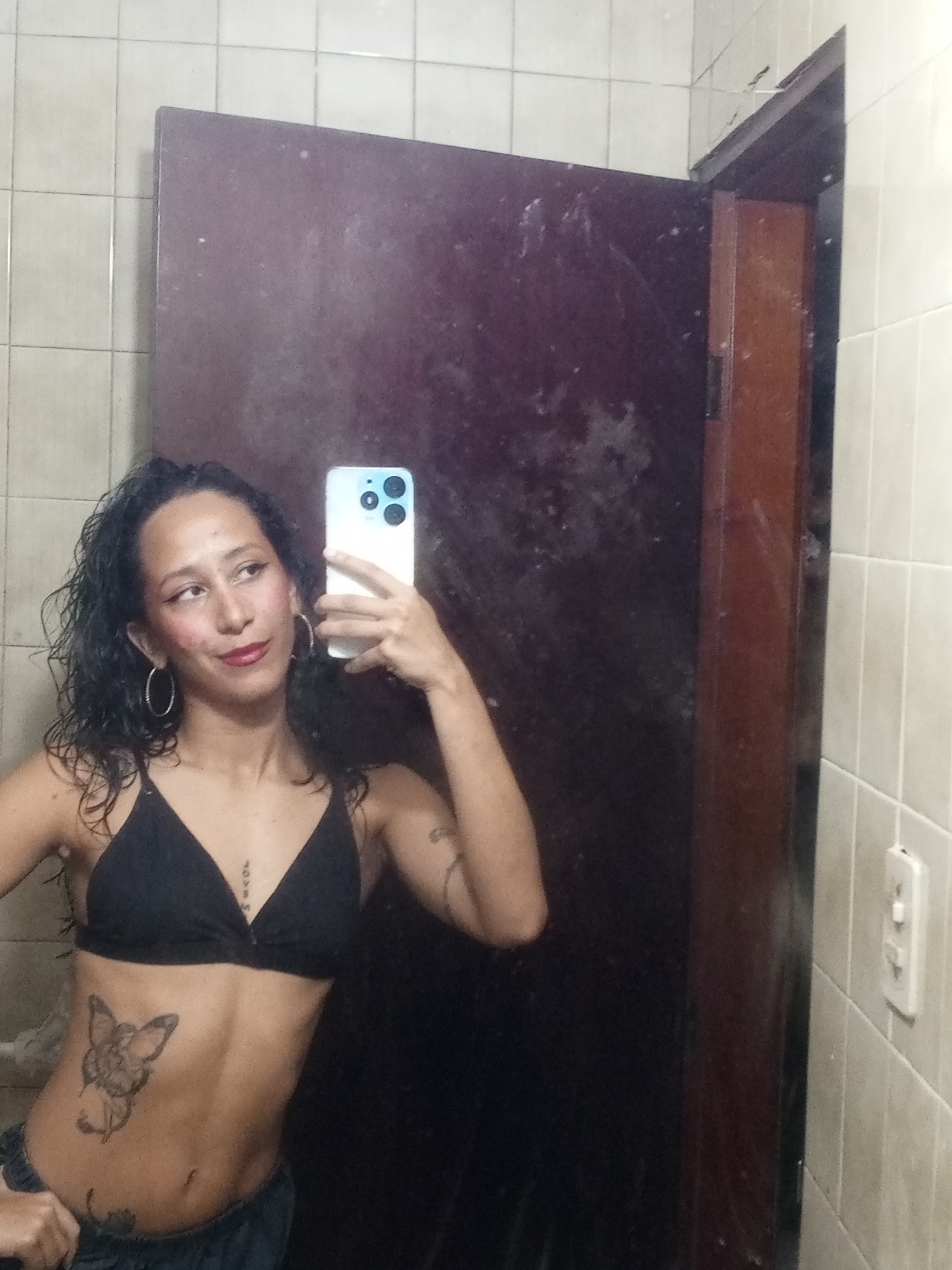 A woman is taking a selfie in a bathroom mirror. She is wearing a black bra and low-rise bottoms. She has curly hair and large hoop earrings. She is holding a phone with a triple-lens camera in her right hand. The background shows a dark-colored door and tiled walls. The light switch is visible on the right wall. The location is likely inside a private residence, but the specific city or country (Porlamar, Venezuela) is not discernible from the image alone.