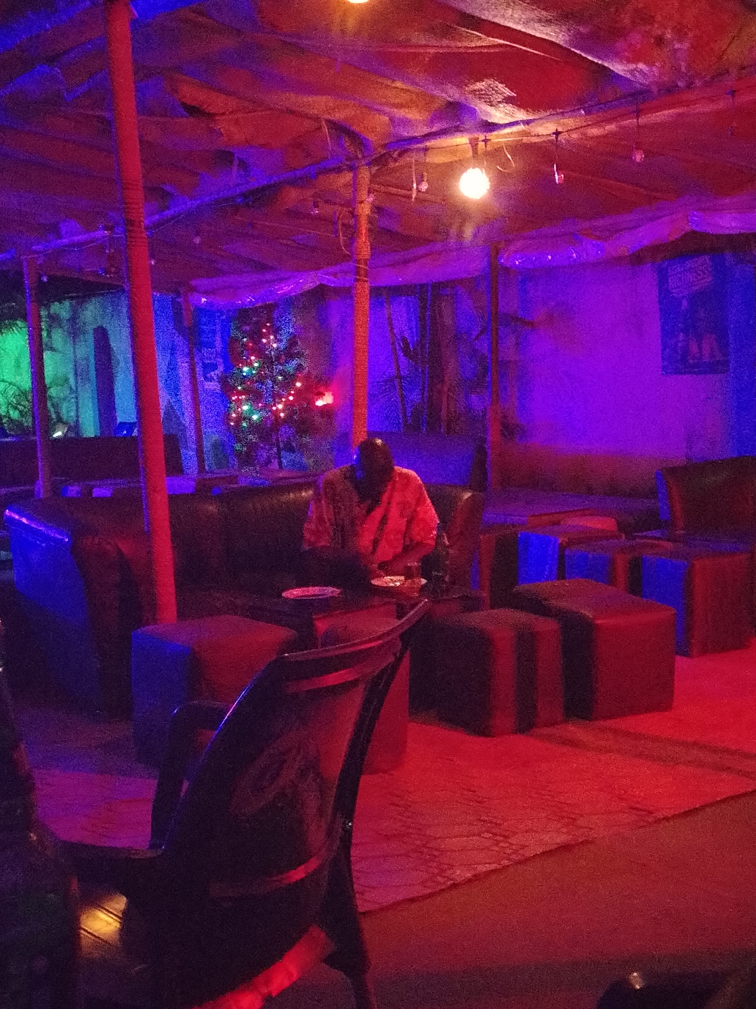 A brightly-lit bar scene with red, blue, and purple lighting. A dark-haired individual sits at a table eating, wearing a light orange, patterned shirt. A Christmas tree with multi-colored lights is visible behind the individual. In the foreground, there is a black chair. In the background, there is a couch, and cube-shaped seating. The room appears to have a makeshift ceiling and is supported by several columns.