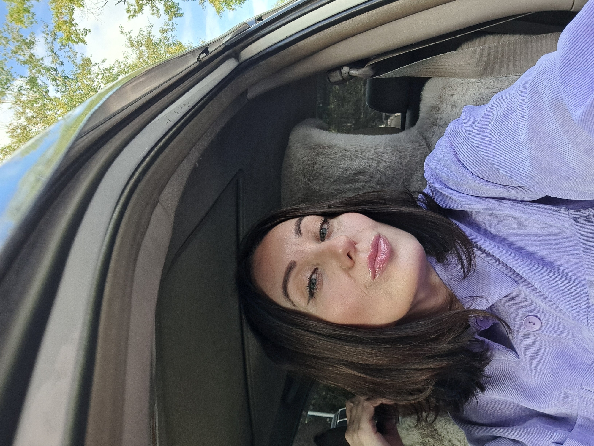 A woman with dark hair and winged eyeliner is taking a selfie in the passenger seat of a car. She is wearing a purple collared shirt and her lips are pursed. The interior of the car is visible, including the fuzzy gray headrest of the driver's seat. The top of the car is open, and green trees and a blue sky are visible above. The image is likely taken during the daytime. There is no information in the image to suggest it is located in Irkutsk, Russia.